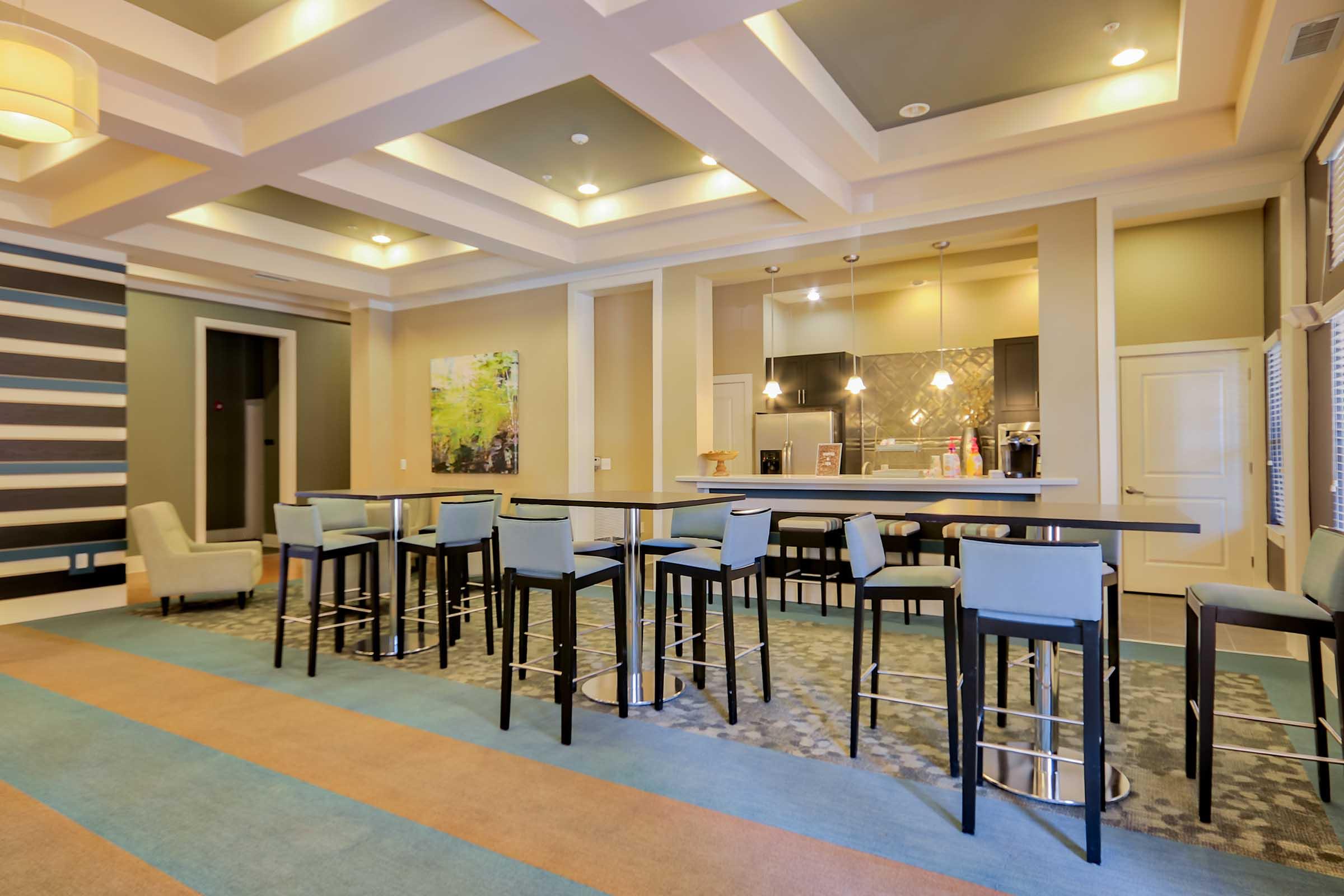 Modern interior of a spacious lounge area featuring a long bar table with high stools, a stylish kitchen in the background, and decorative walls. The soft color palette includes greens, blues, and neutral tones, creating a welcoming atmosphere. Large windows allow natural light to fill the space.