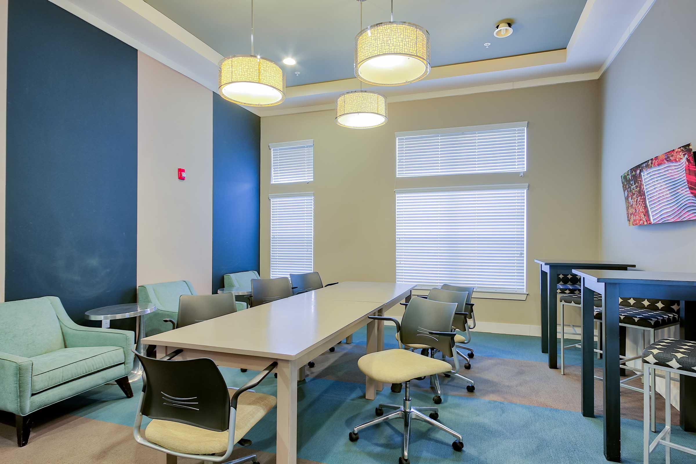 A modern conference room featuring a large rectangular table surrounded by black office chairs. In the background, there are teal armchairs and side tables. Three hanging light fixtures illuminate the space, which has large windows with blinds. A decorative wall art piece is visible on the right.