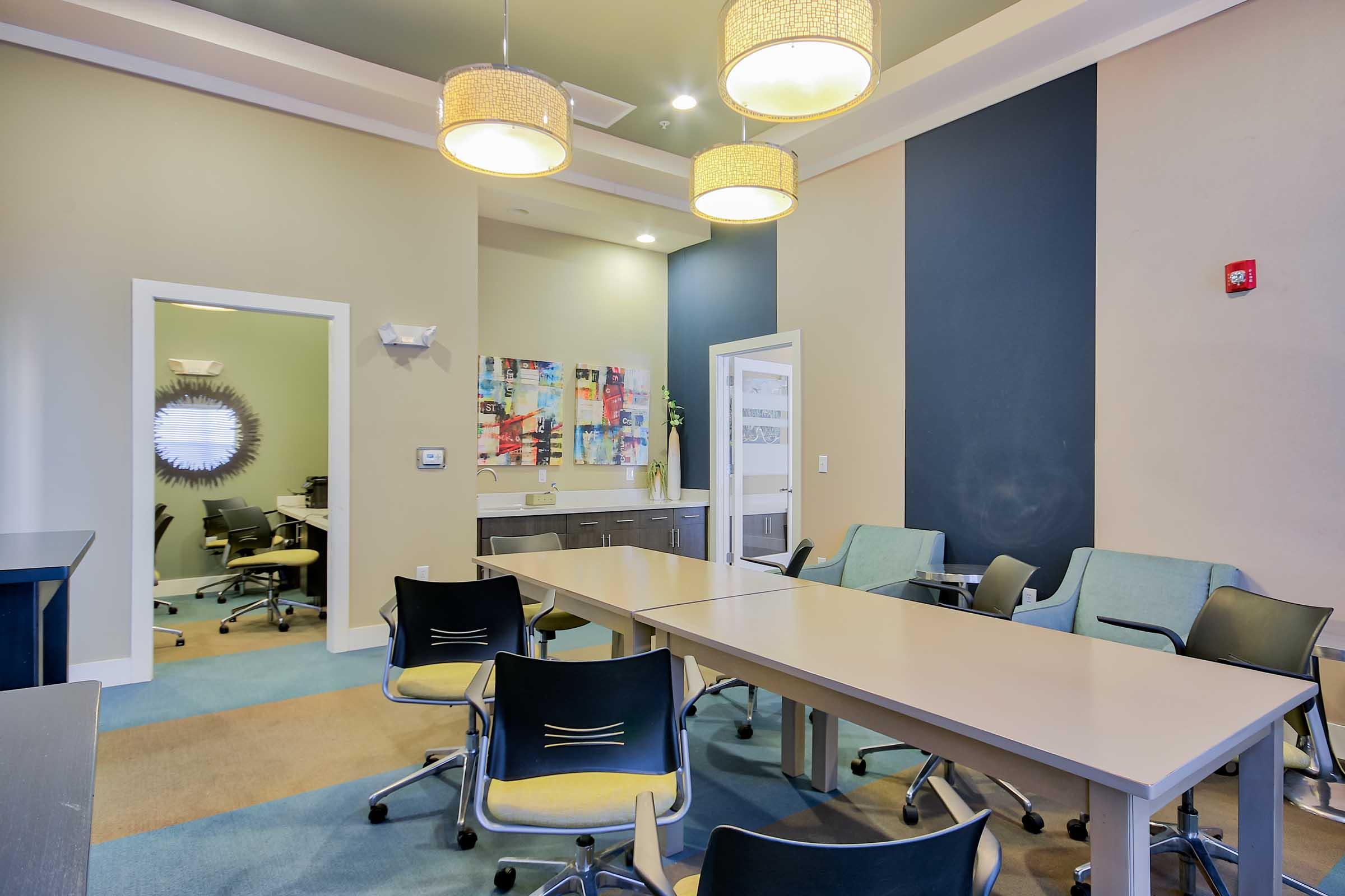 A modern conference room featuring a long table with rolling chairs, surrounded by neutral-colored walls. Two pendant lights hang from the ceiling, and there's a kitchenette with cabinets and a colorful wall art piece. In the background, a glimpse of another room with office chairs can be seen.