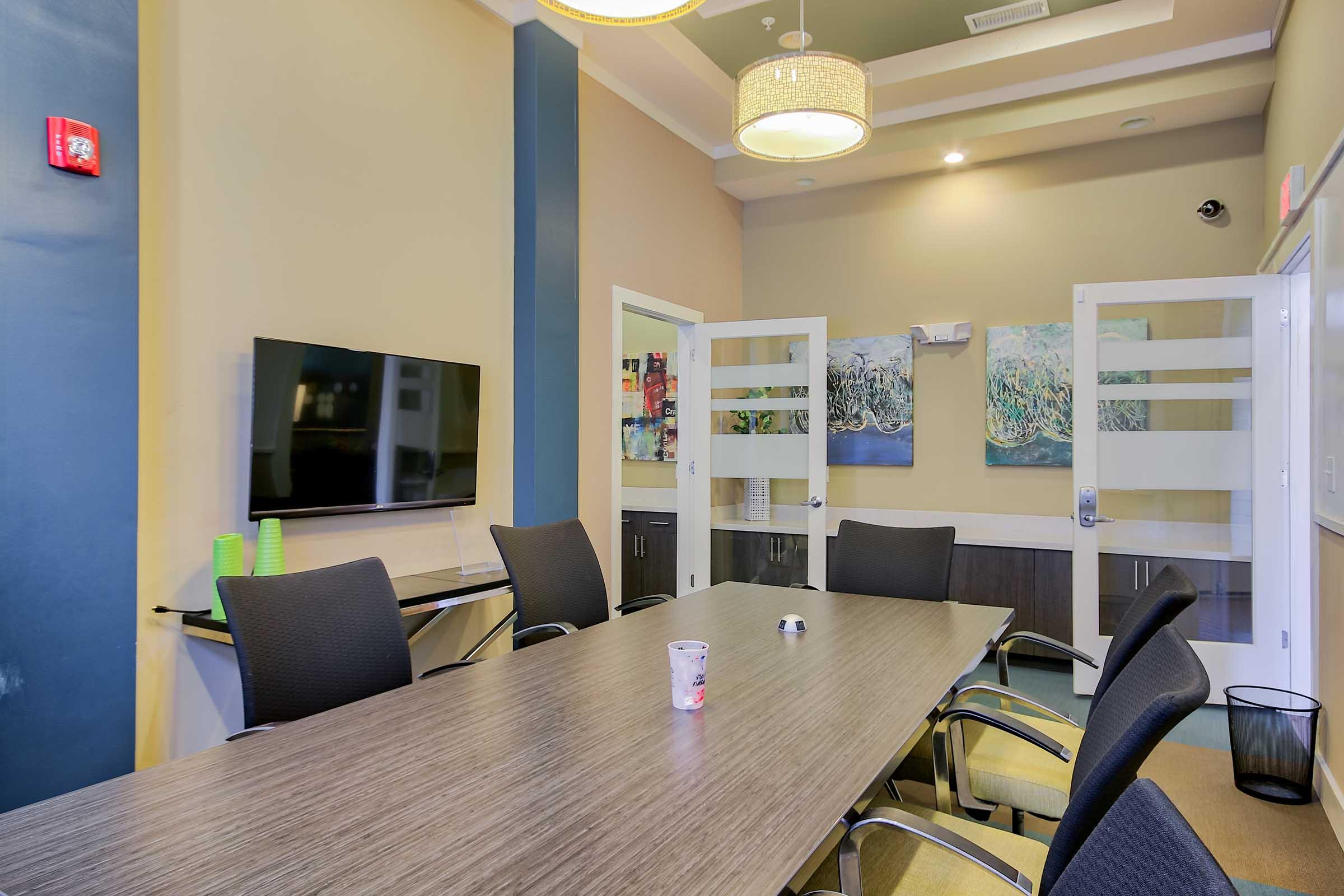 A modern conference room featuring a long wooden table surrounded by black chairs. There is a wall-mounted television on one side, and two glass doors leading to another area. The walls are painted in soft colors, with abstract artwork displayed. Ambient lighting is provided by a ceiling fixture.