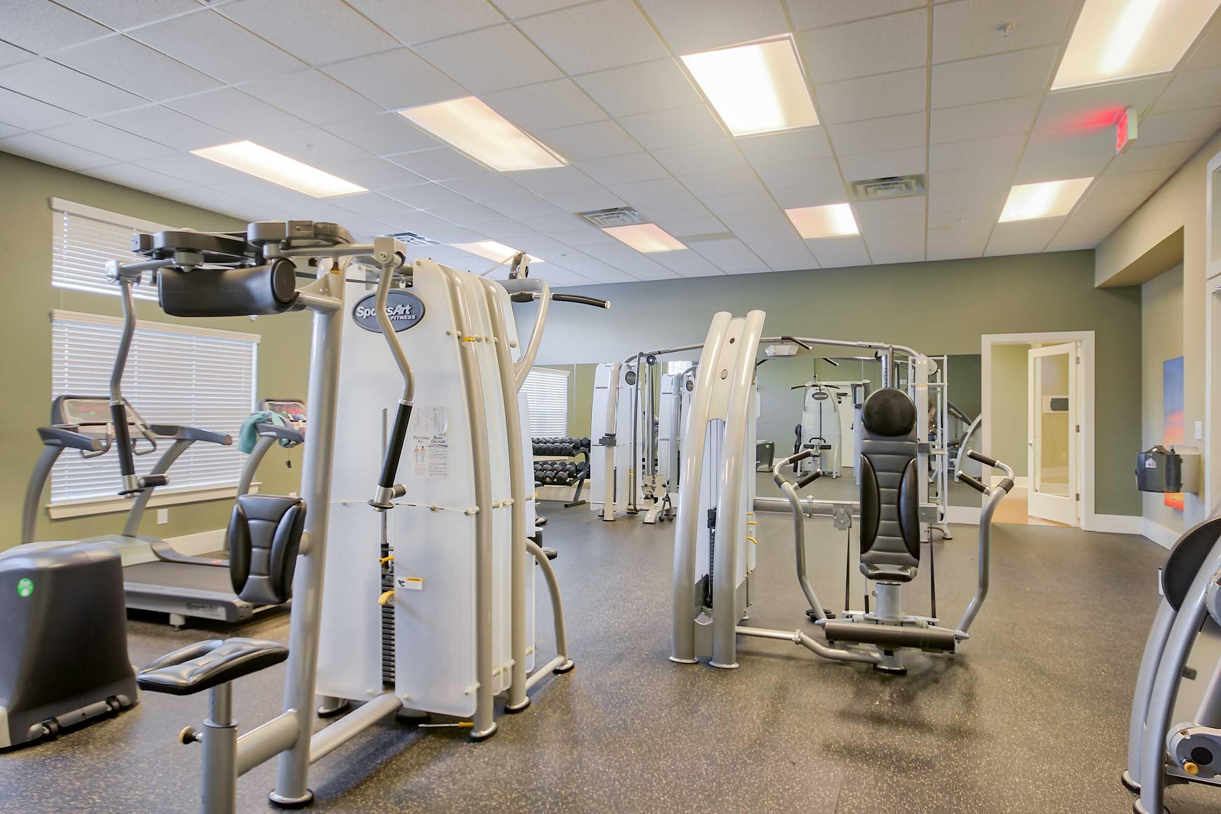 A spacious gym interior featuring a variety of fitness equipment, including weight machines and cardio machines. The room has bright overhead lighting, large windows with natural light, and a clean, modern aesthetic, ideal for workouts.