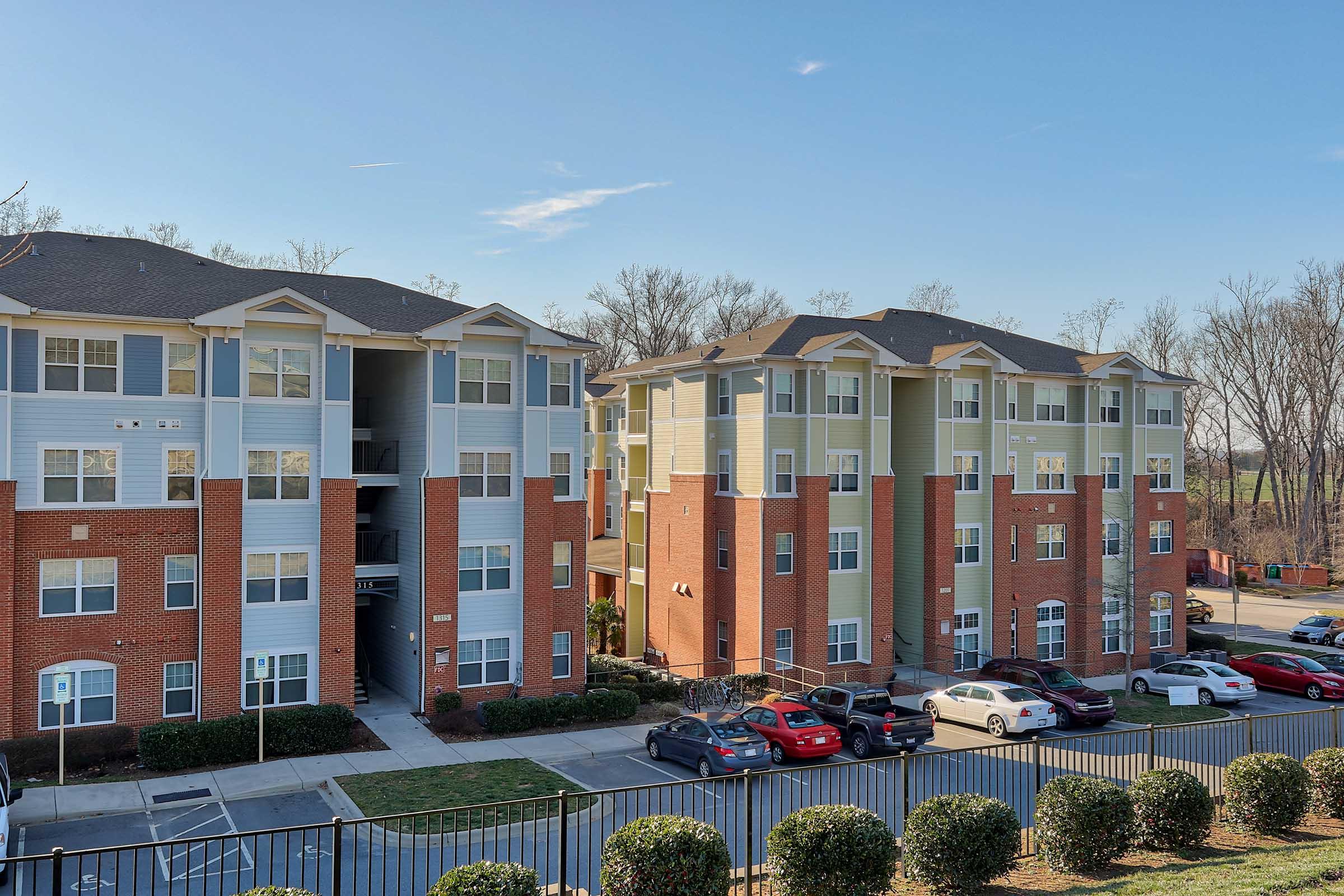 Two modern brick apartment buildings with multiple floors, featuring balconies and windows. The foreground shows neatly landscaped bushes and a parking area with parked cars. In the background, there are trees and a clear blue sky.