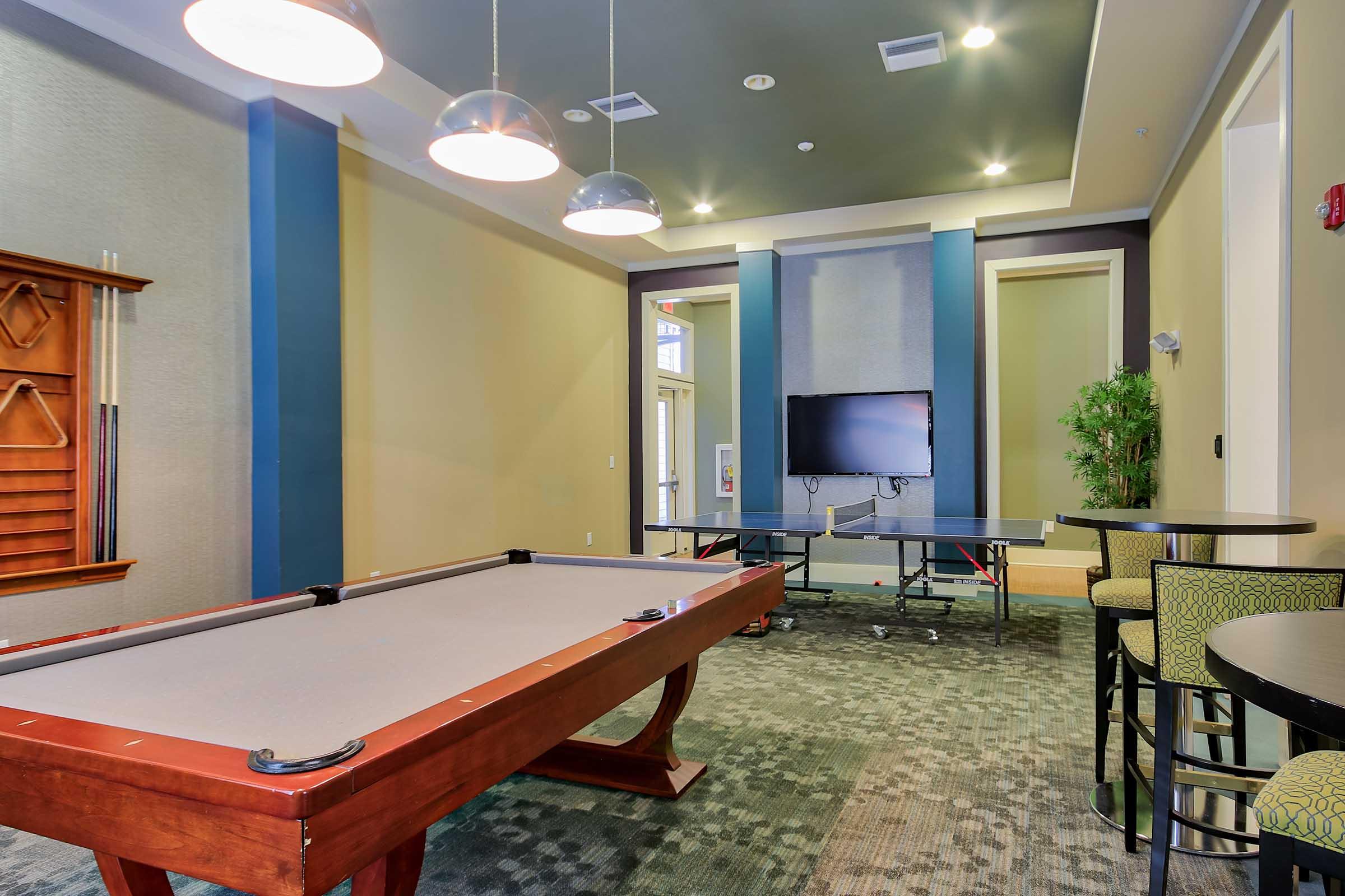 A recreational room featuring a billiards table, a television on the wall, and a ping pong table. The space has modern lighting fixtures hanging from the ceiling and a mix of green and beige decor on the walls and floor. There are high-top tables and stools for seating.