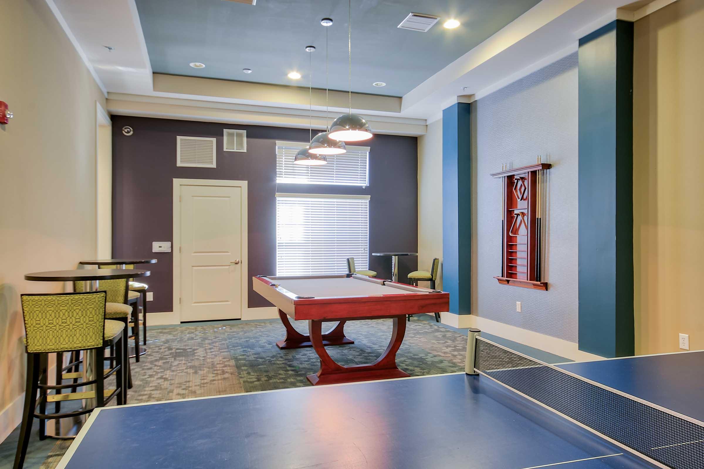 A recreation room featuring a red pool table, a blue ping pong table, and high-top seating. Large windows allow natural light, and stylish lighting hangs above the pool table, creating a vibrant, inviting space for leisure and socializing.