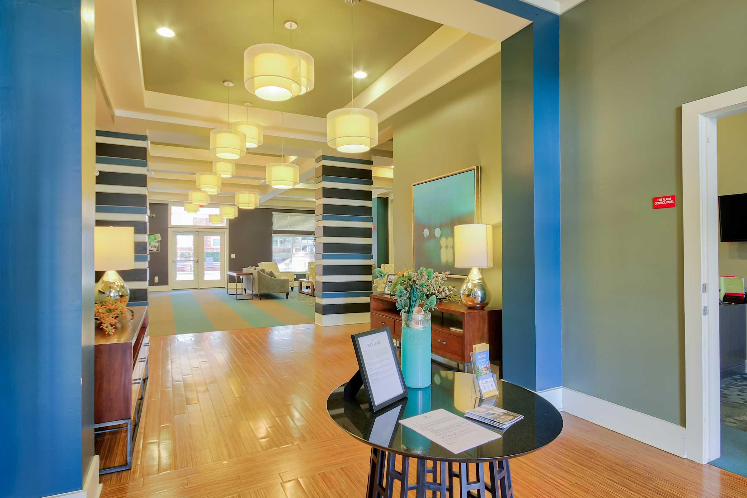 Modern lobby interior featuring stylish lighting fixtures, hardwood flooring, and contemporary decor. A round table with informational materials sits in the foreground, while seating areas and artwork create an inviting atmosphere in the background.