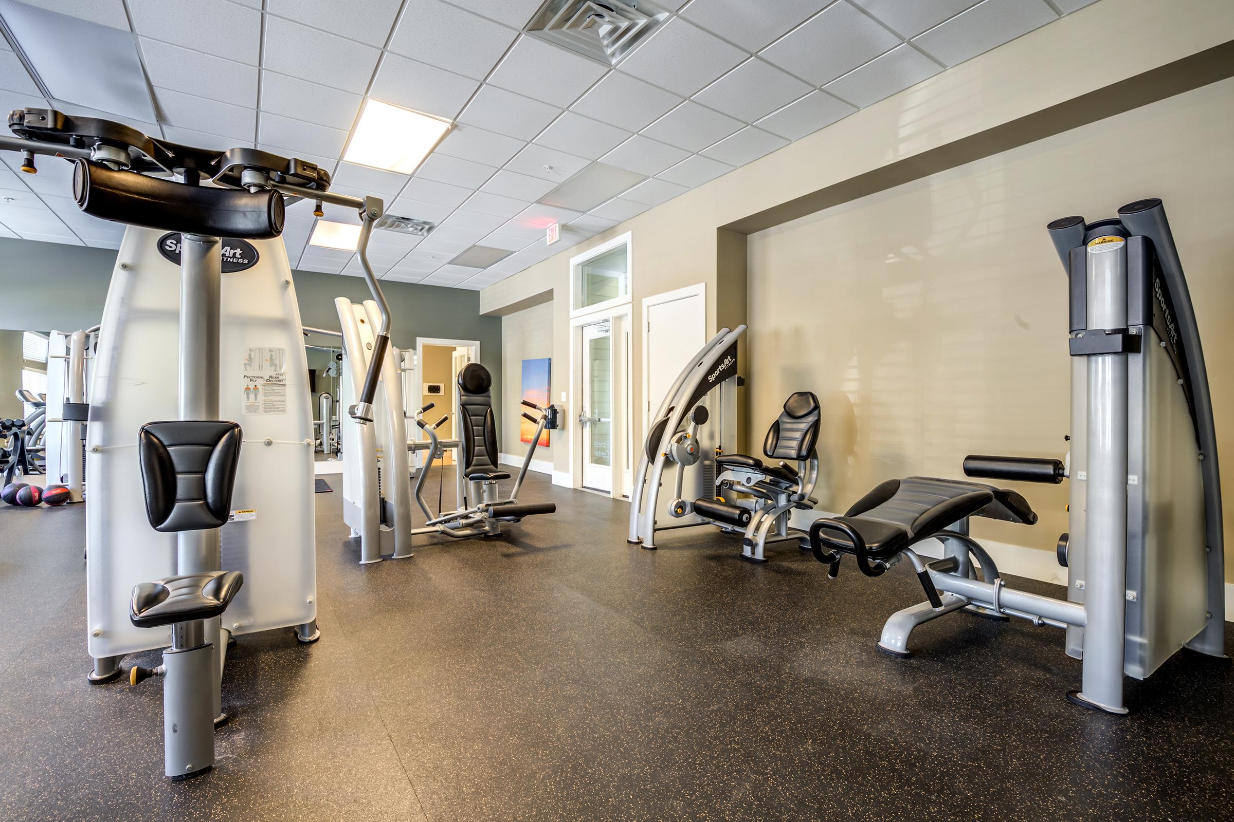 A modern gym interior featuring various strength training equipment, including multi-station machines, a leg press, and a bench. The floor has a rubberized surface, and large windows allow natural light to fill the space. Wall colors are neutral, creating a clean and inviting environment for workouts.