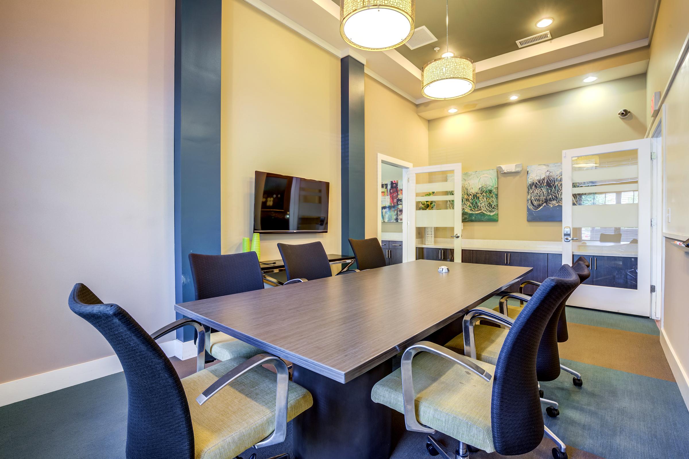 A modern conference room featuring a large rectangular table surrounded by ergonomic chairs. The room has a flat-screen TV mounted on the wall, a whiteboard, and decorative artwork. Soft lighting from overhead fixtures enhances the welcoming atmosphere, while neutral paint colors create a professional environment.