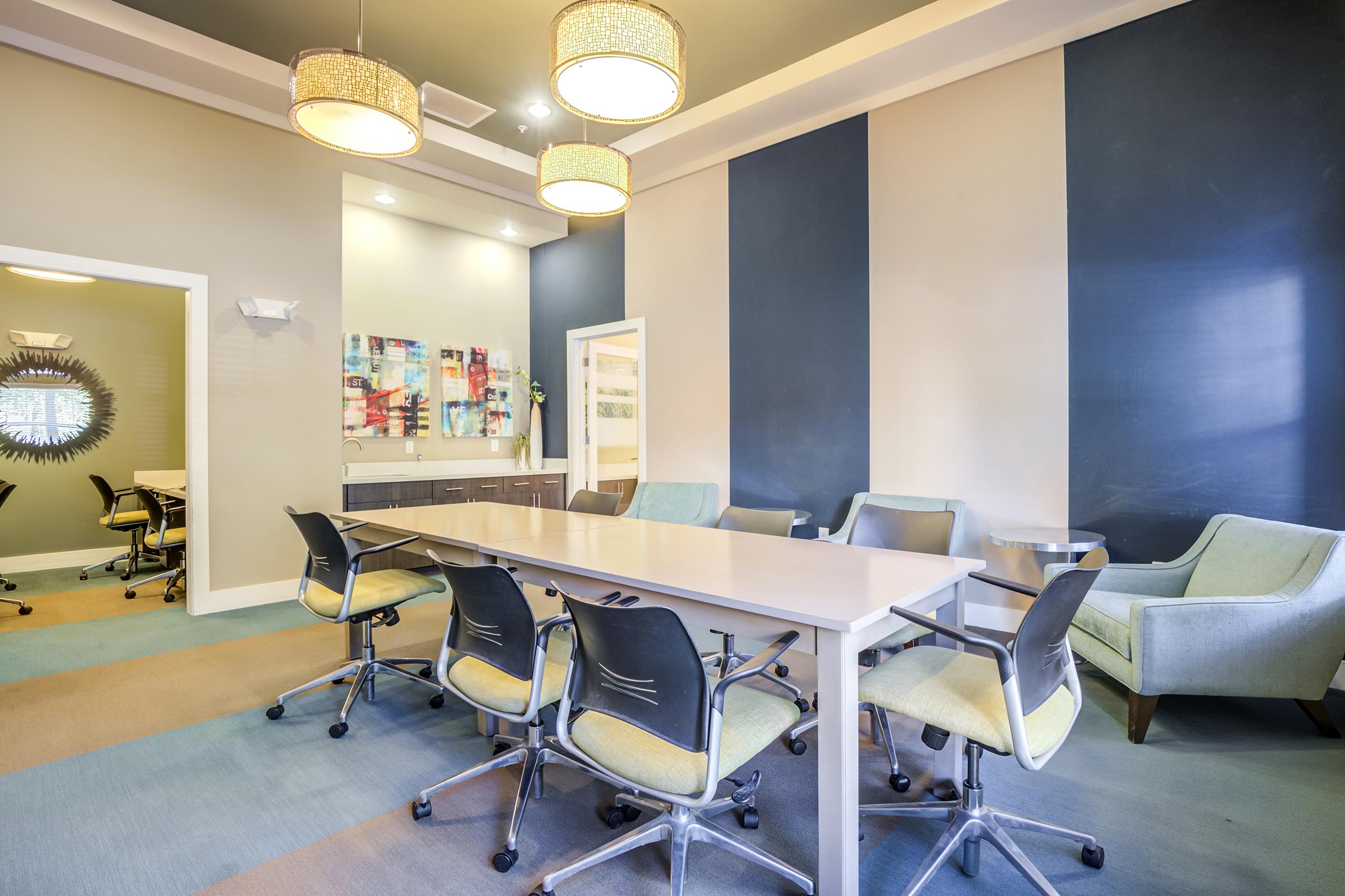 Modern conference room with a long table surrounded by ergonomic chairs. The walls feature a two-tone paint design, and the room is illuminated by stylish pendant lights. In the background, there's a kitchenette area and a cozy seating space with armchairs.