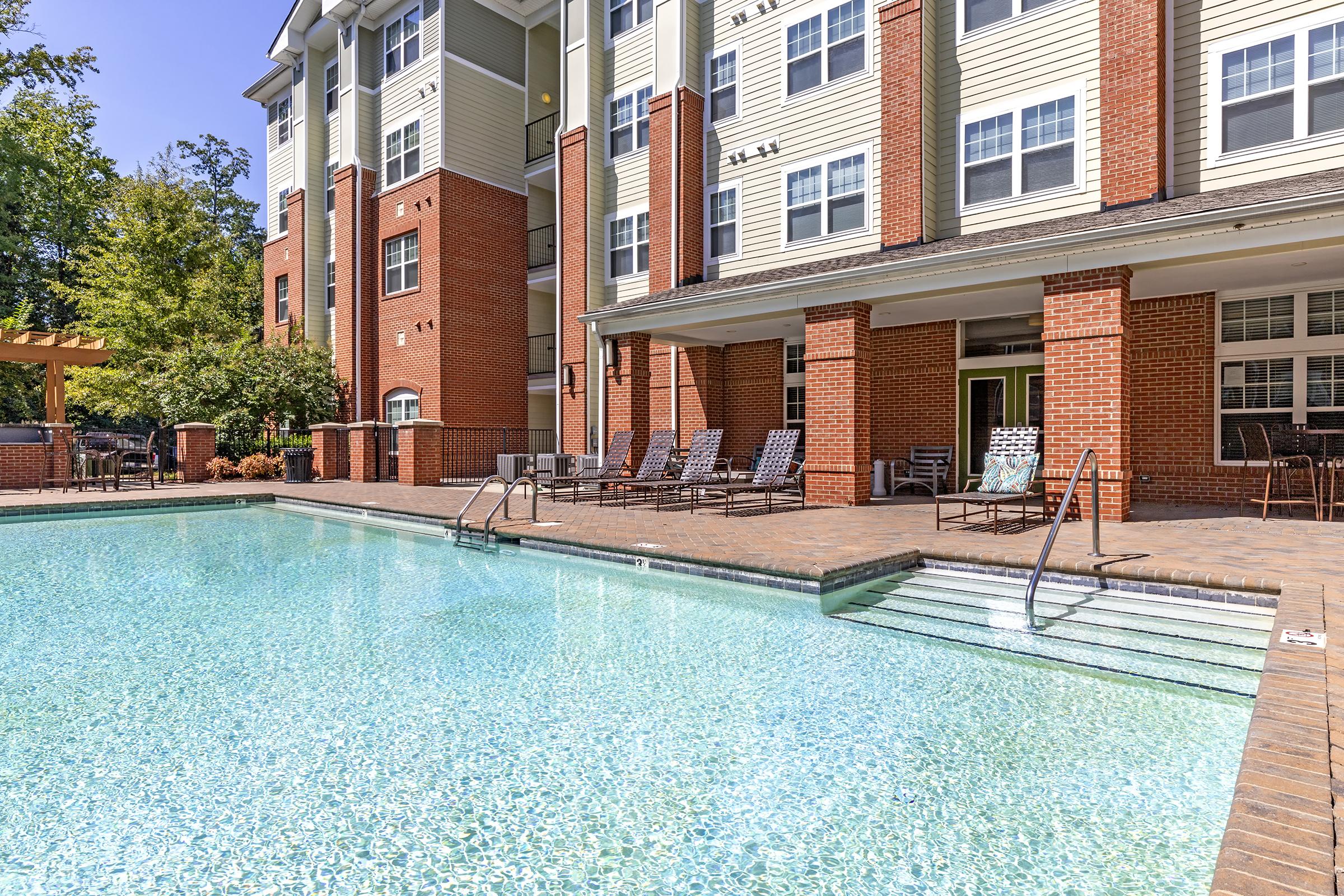 A clear blue swimming pool with tiled steps, surrounded by lounge chairs. The pool area is adjacent to a multi-story brick building with large windows and balconies. Lush greenery is visible in the background, indicating a sunny day.