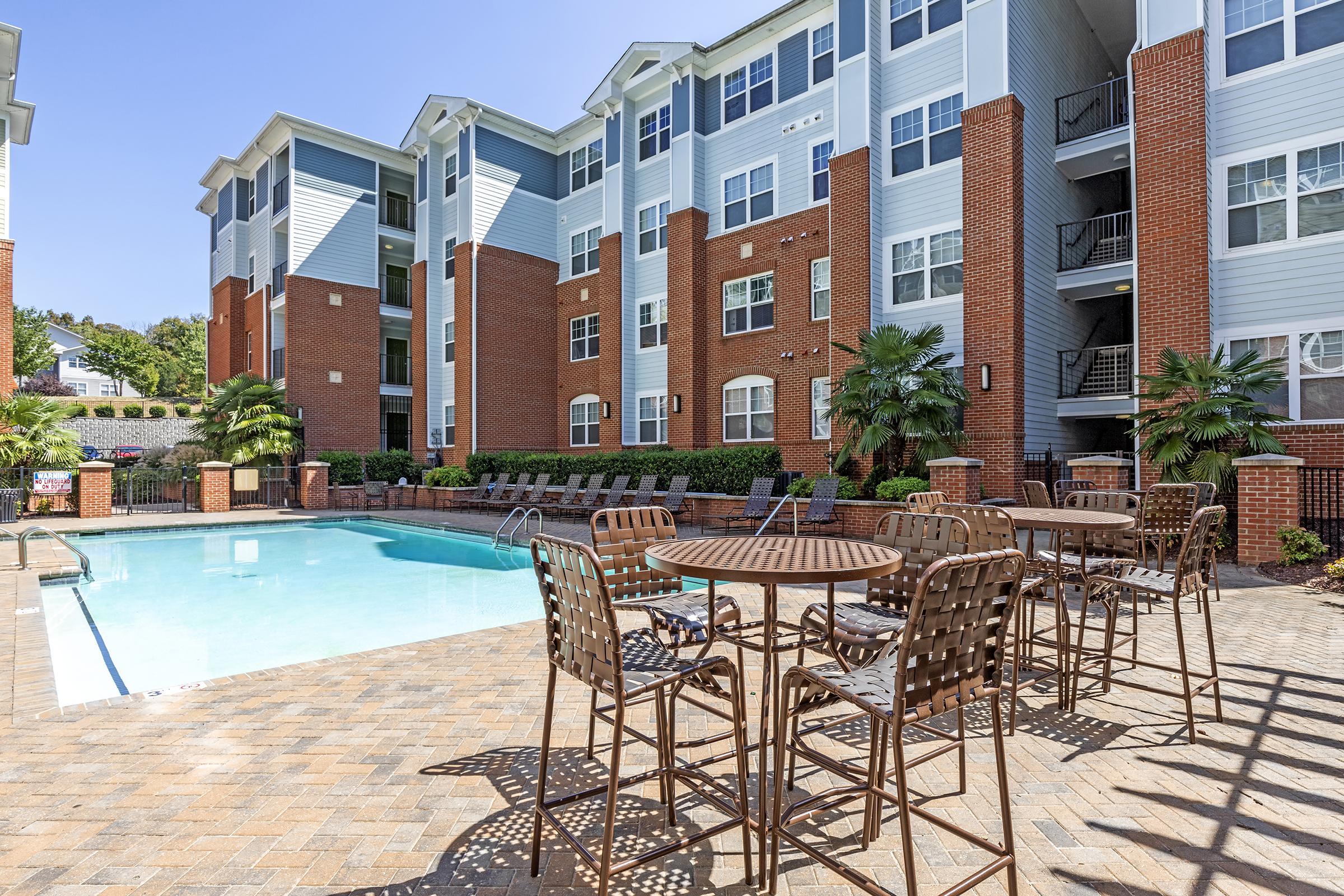 A residential apartment complex featuring a swimming pool surrounded by lounge chairs and tables. The buildings are modern, with brick exteriors and large windows, set against a bright blue sky. Lush greenery adds to the inviting atmosphere of the outdoor space.