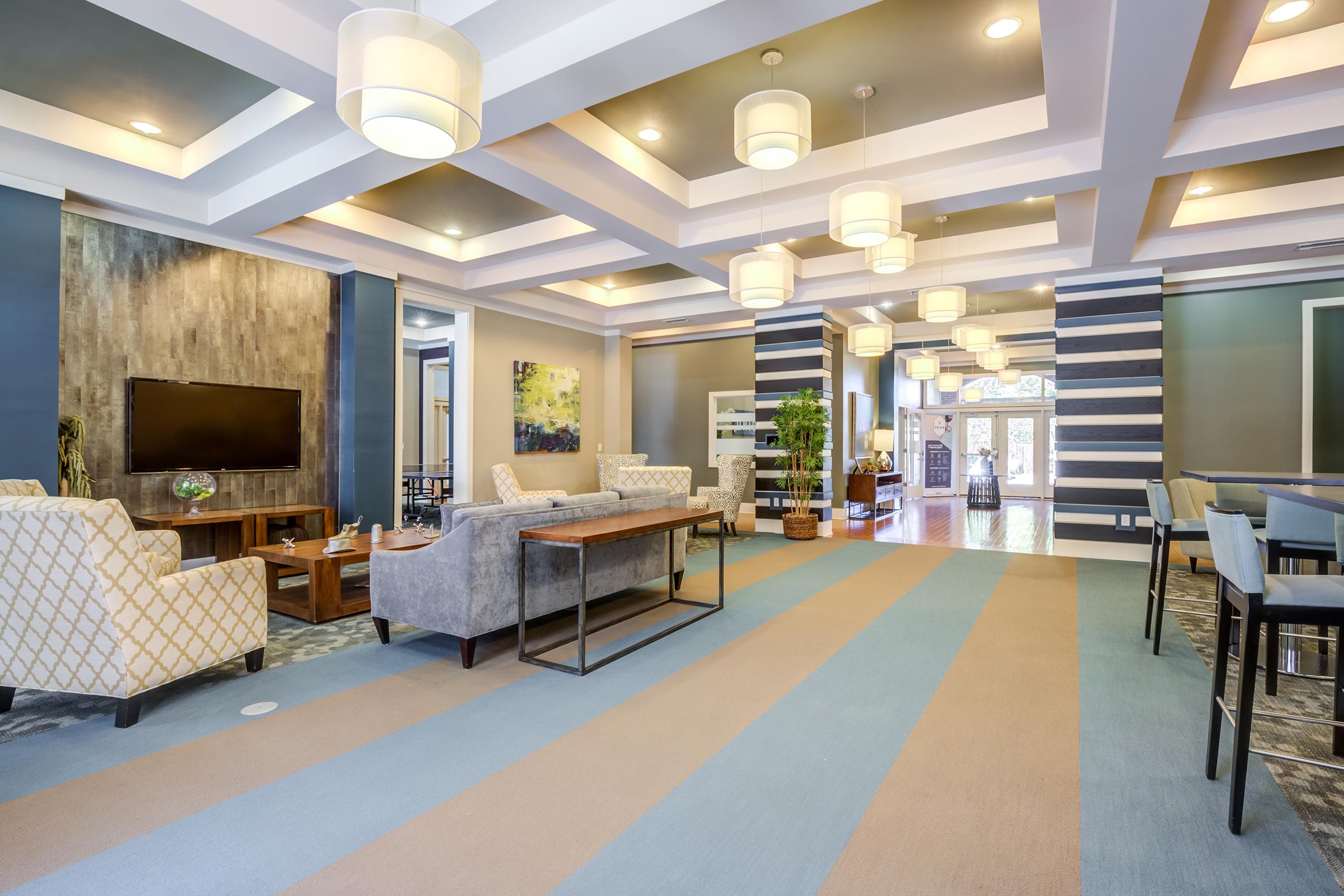 A modern lobby area featuring stylish furniture, including patterned armchairs and a coffee table. The ceiling is adorned with decorative lights, and large windows allow natural light to fill the space. A television is mounted on the wall, and potted plants add a touch of greenery to the contemporary design.