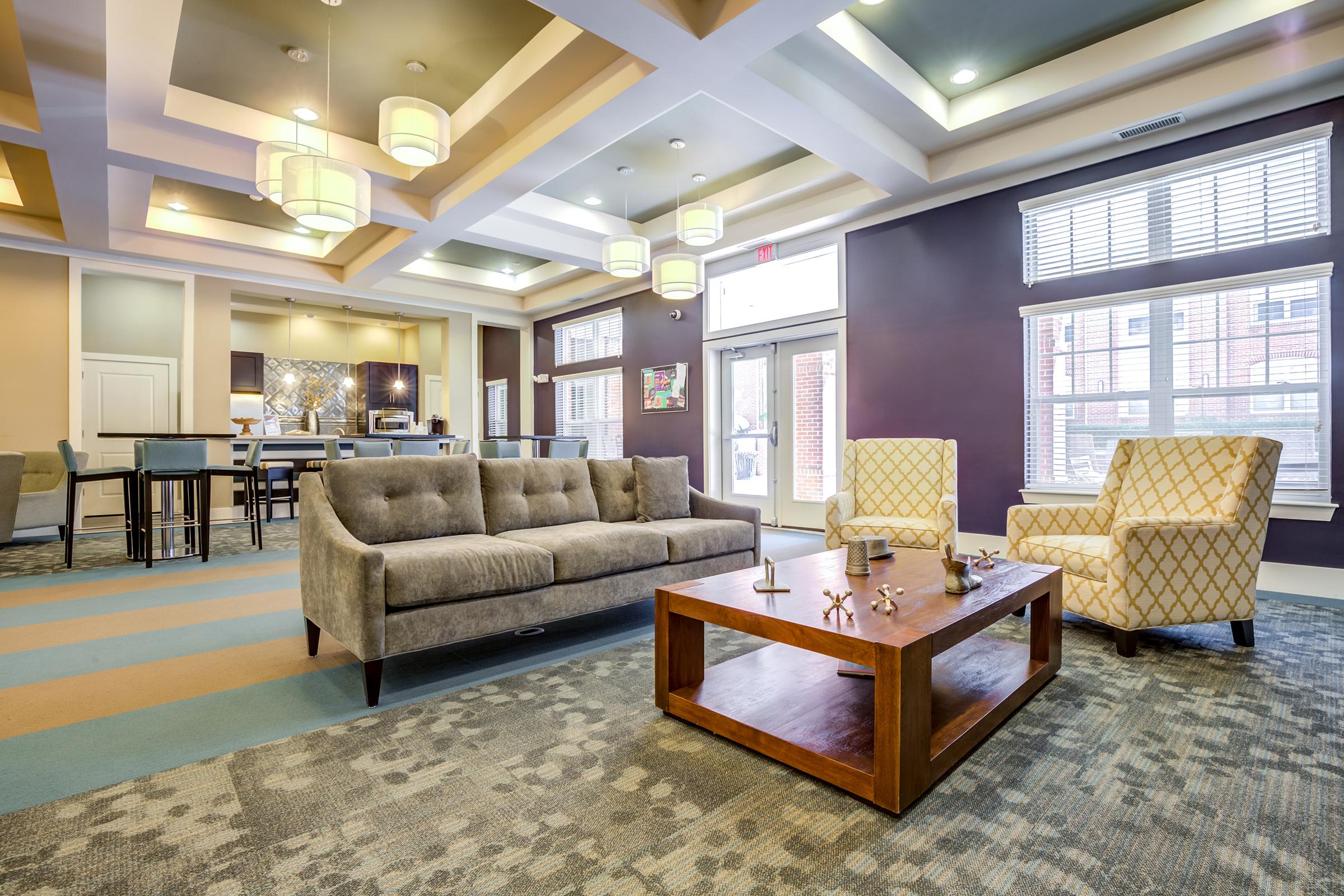 A modern living room featuring a stylish gray sofa, two patterned armchairs, and a wooden coffee table. The space has large windows allowing natural light, with a ceiling featuring decorative lighting and a contemporary color palette. In the background, a kitchen can be seen.