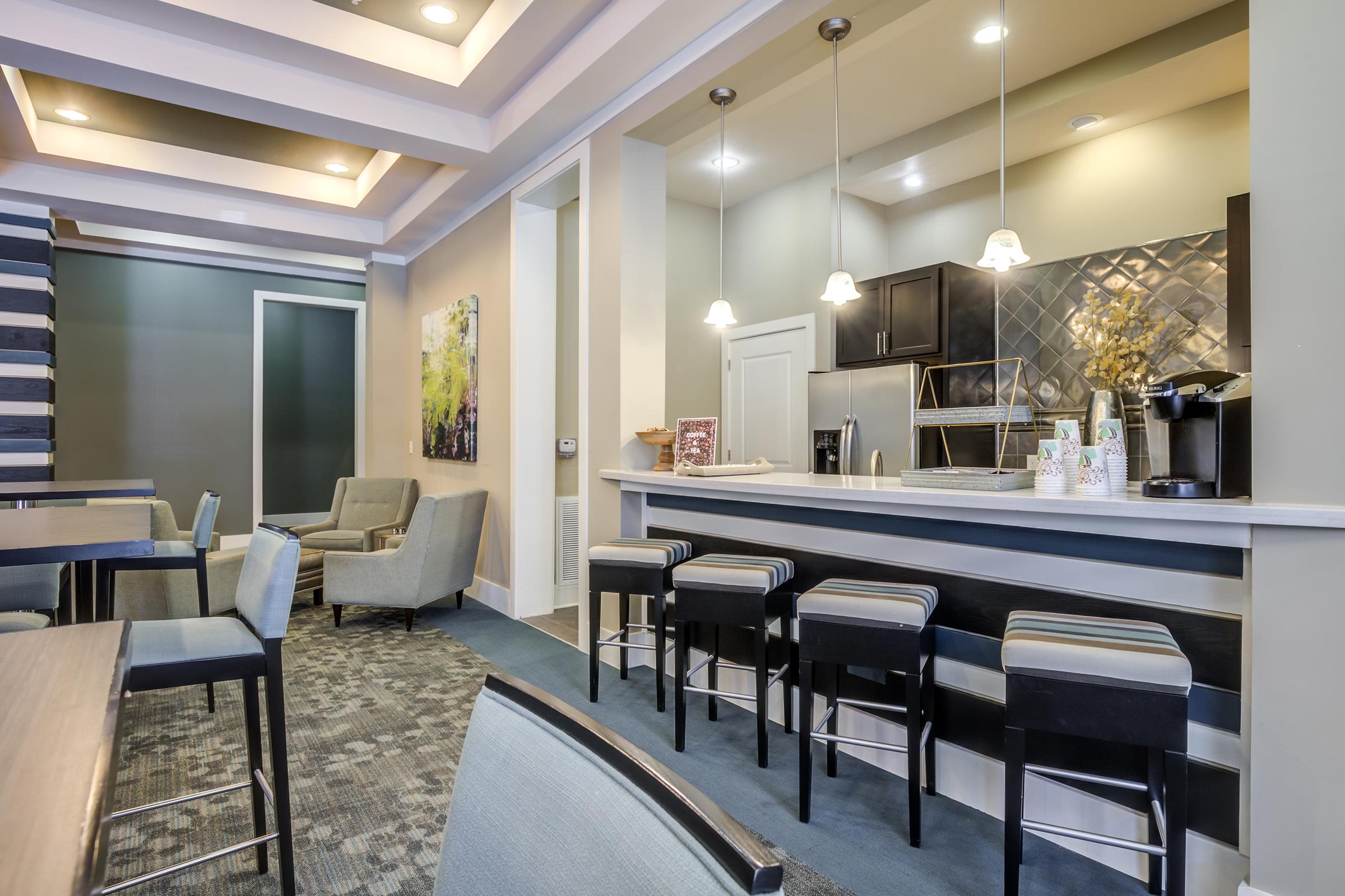 A modern, stylish kitchen and dining area featuring a sleek countertop with bar stools, a coffee maker, and cabinetry. The space is well-lit with recessed lighting, complemented by contemporary seating and decorative artwork on the walls, creating a welcoming atmosphere for socializing and dining.