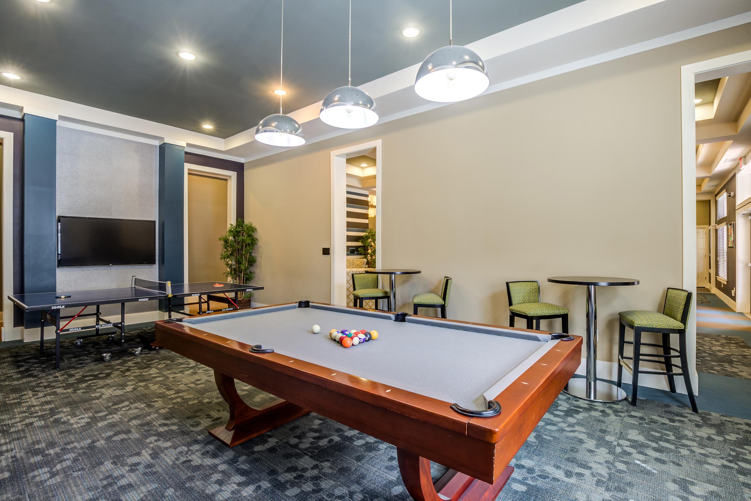A well-lit game room featuring a pool table with balls set for play. Adjacent to the table are two small round tables with chairs, and a large flat-screen TV mounted on the wall. The walls are painted in neutral tones, enhancing the modern and inviting atmosphere of the space.