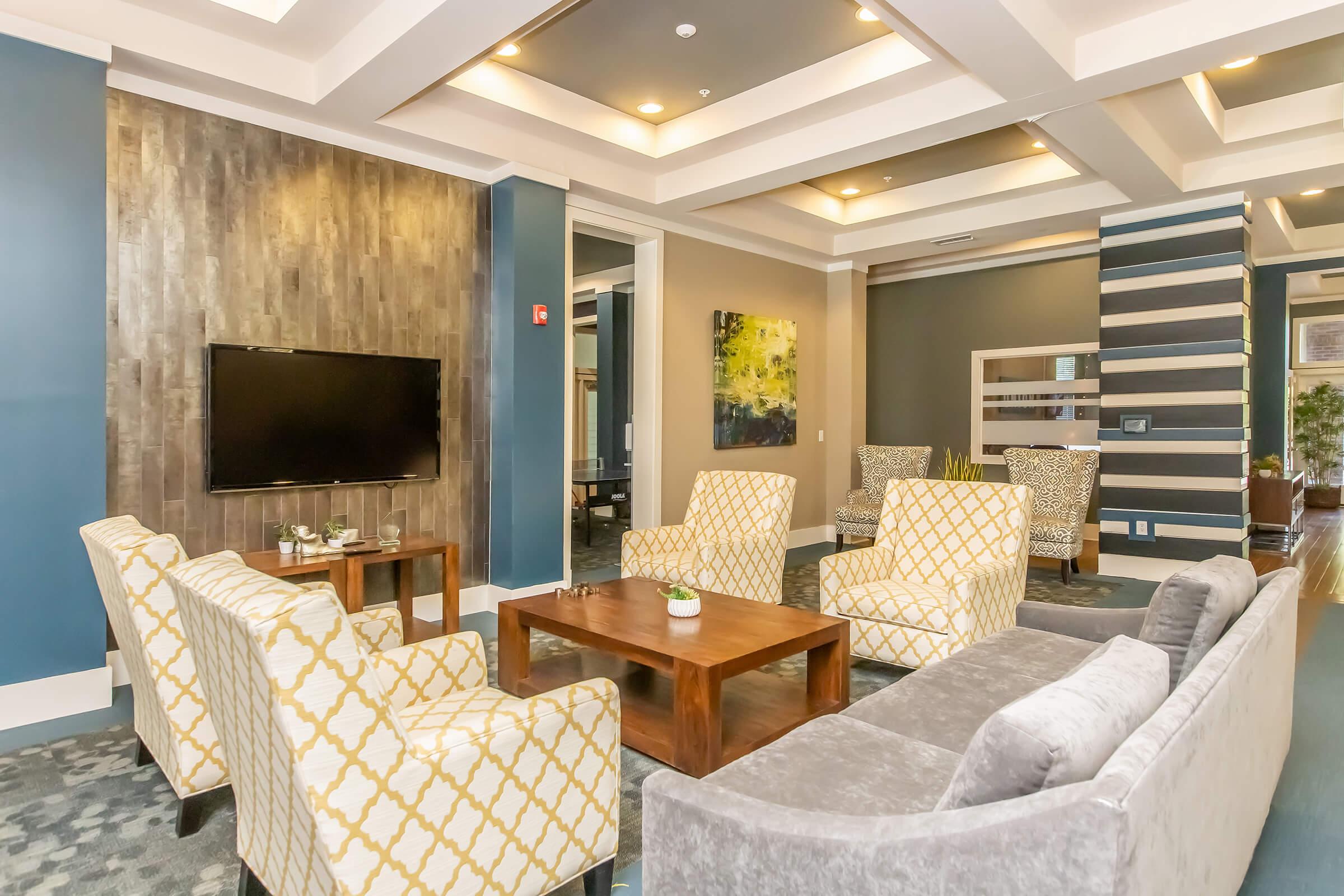 A cozy lounge area featuring a modern design with comfortable seating, including yellow patterned armchairs and a grey sofa. There's a wooden coffee table in the center, and a flat-screen TV mounted on the wall. The walls are painted in blue tones with decorative artwork, creating a welcoming atmosphere.