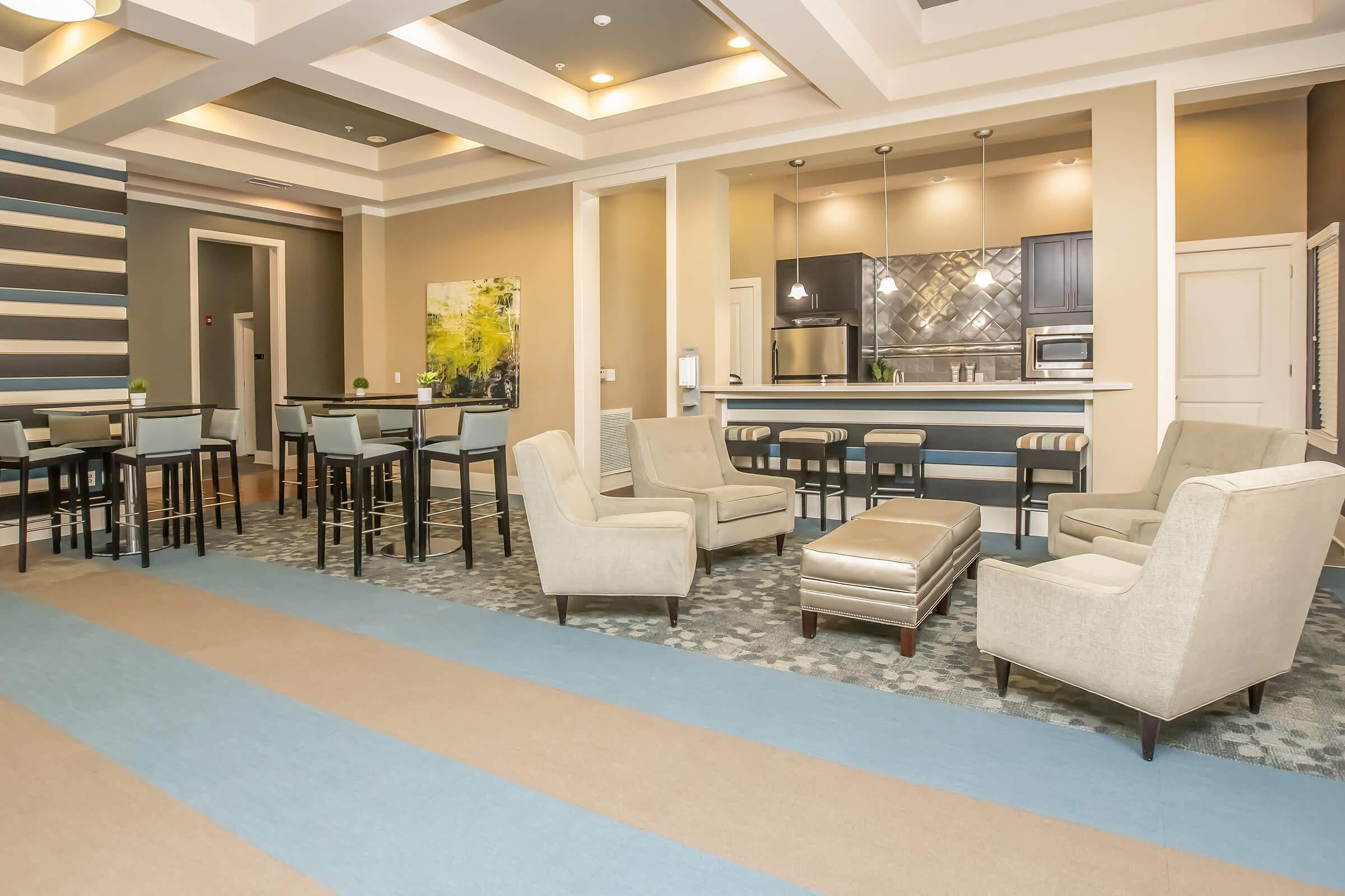 A modern lounge area featuring a combination of light and dark furniture. Cozy seating arrangements with armchairs and a tufted ottoman are set on a patterned carpet. A bar-height table with chairs is visible, along with a kitchenette in the background, showcasing stylish cabinetry and tile work.