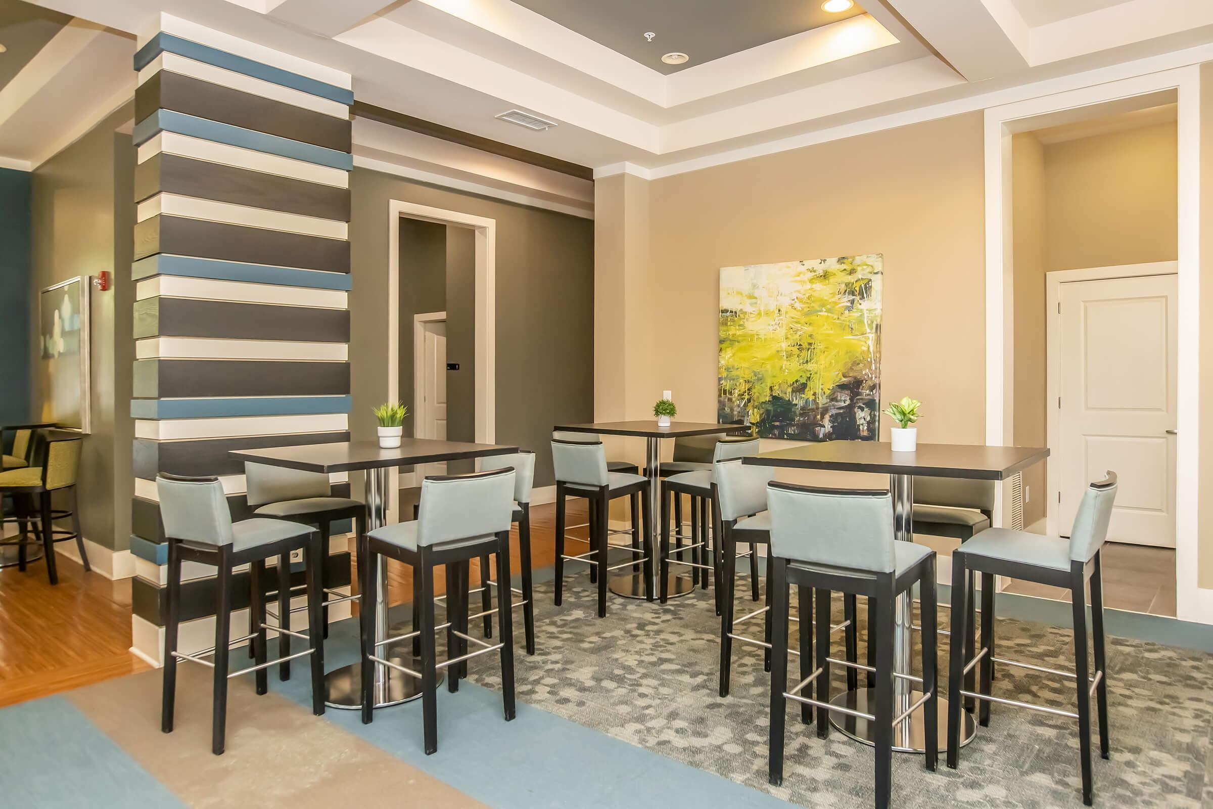 A modern, well-lit dining area featuring tall black tables with light blue chairs. The space includes a decorative plant on each table and has contemporary wall art with bright colors. The background showcases neutral walls and a multi-striped pillar, creating a stylish ambiance.