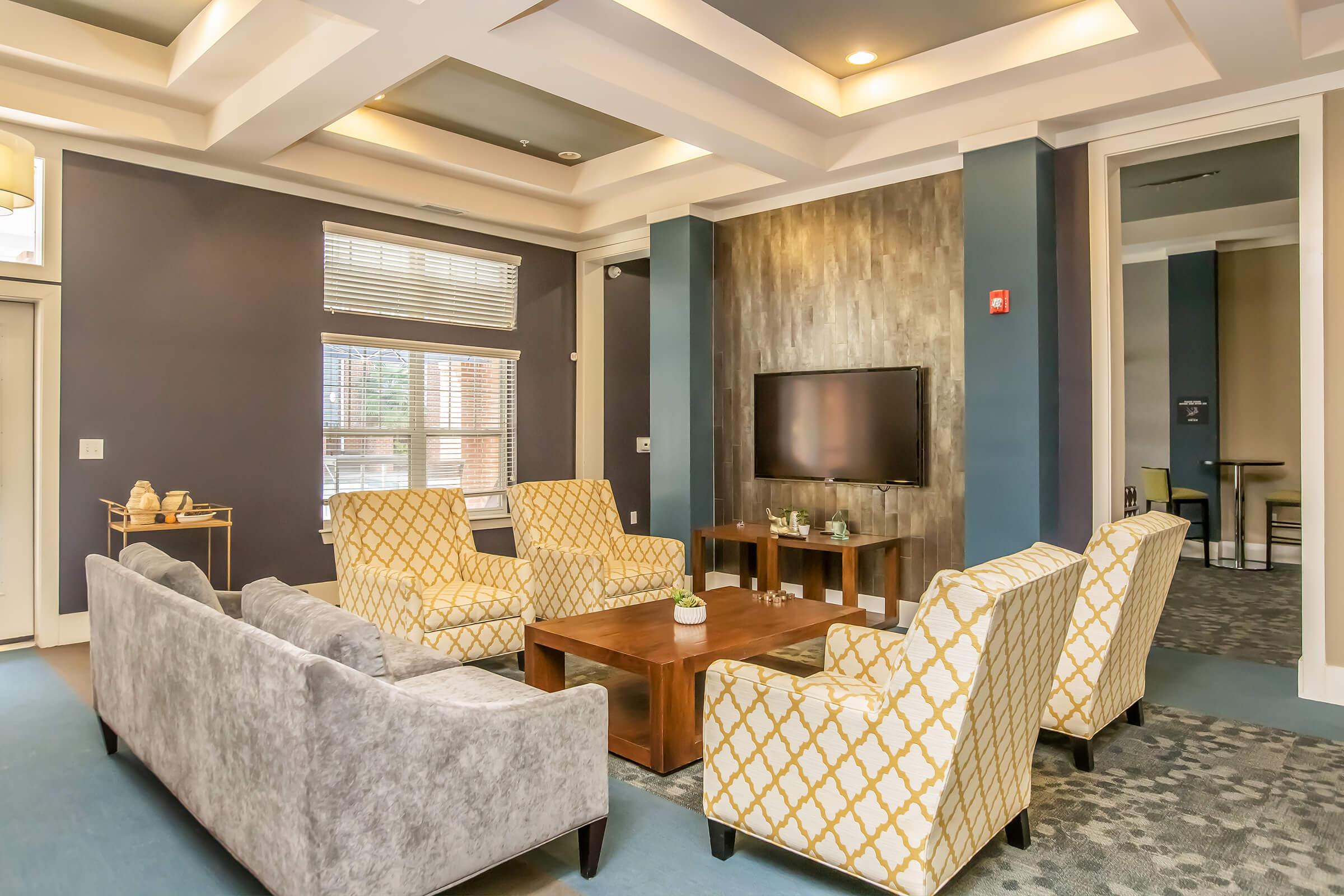 A cozy lounge area featuring a gray sofa and four patterned chairs surrounding a wooden coffee table. A flat-screen TV is mounted on the wall, and large windows provide natural light. The walls are painted in a deep blue with decorative molding, creating a welcoming atmosphere.