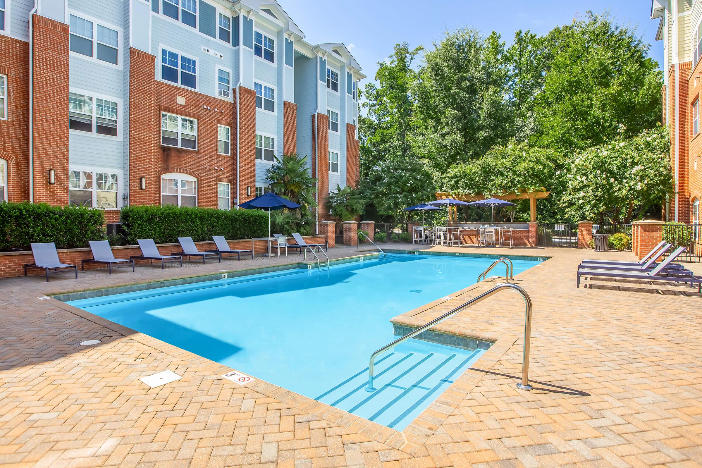 A clear blue swimming pool surrounded by brick pavement, featuring lounge chairs and shaded areas under umbrellas. Lush greenery and trees line the background, while modern apartment buildings are visible nearby, creating a relaxing outdoor atmosphere.