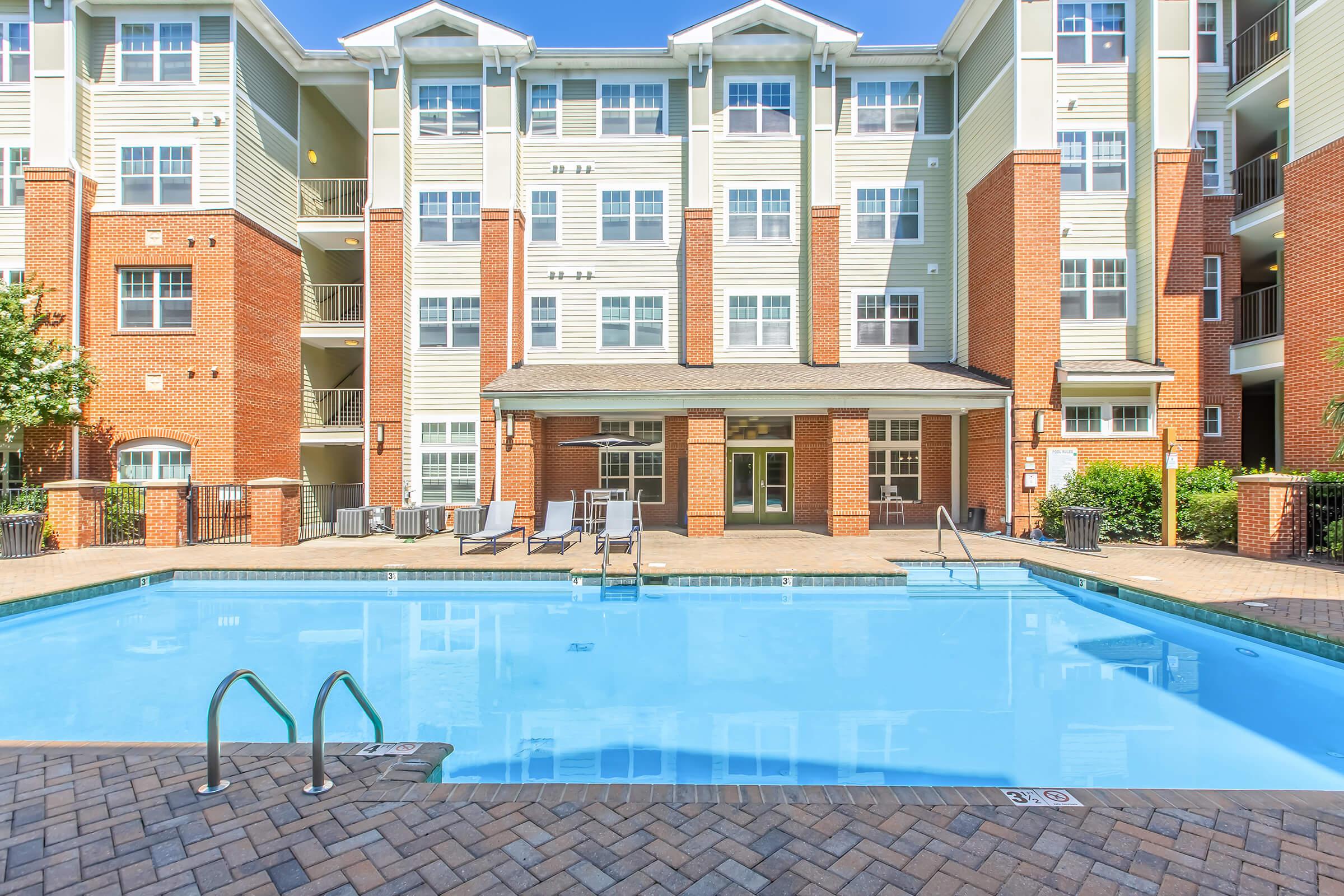 A clear blue swimming pool surrounded by a brick patio. In the background, a multi-story apartment building features light-colored siding and red brick accents. Lounge chairs are positioned nearby, and lush greenery adds to the inviting atmosphere. Bright sunlight highlights the scene.