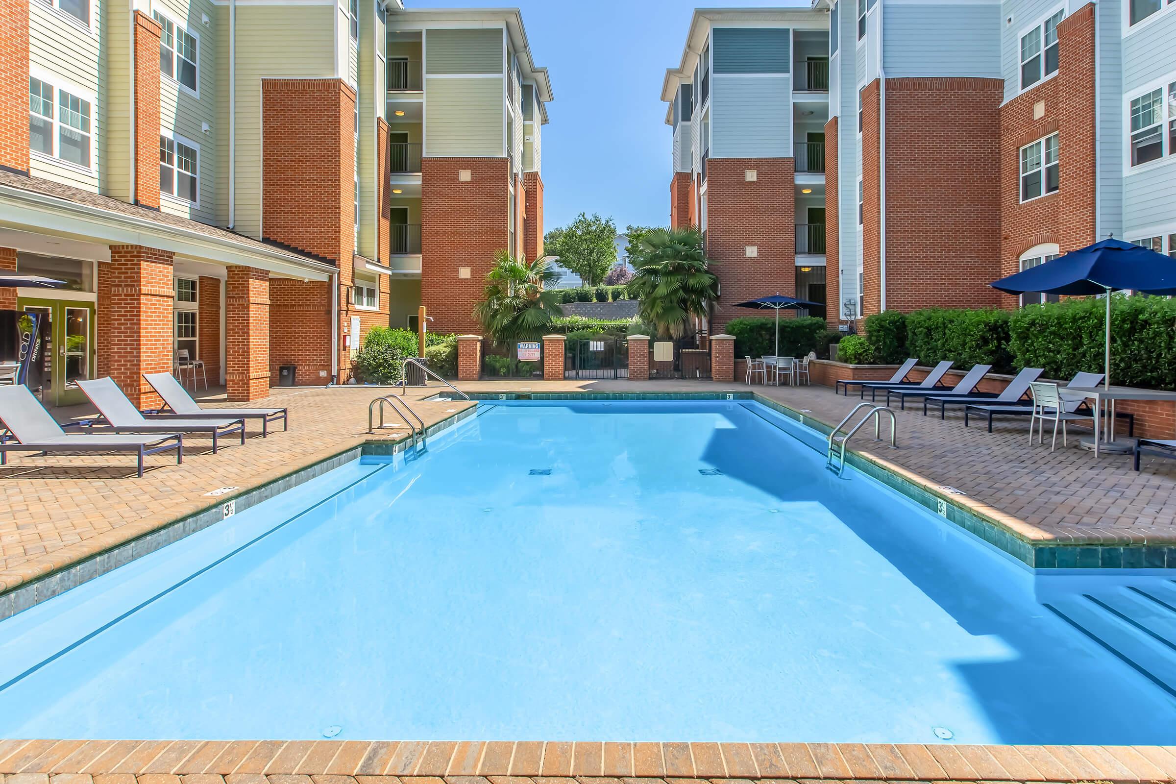 A clear blue swimming pool surrounded by brick-paved decking and lounge chairs. Tall, modern apartment buildings in the background, featuring balconies and green landscaping. Sunlight illuminates the scene, creating a relaxing atmosphere.