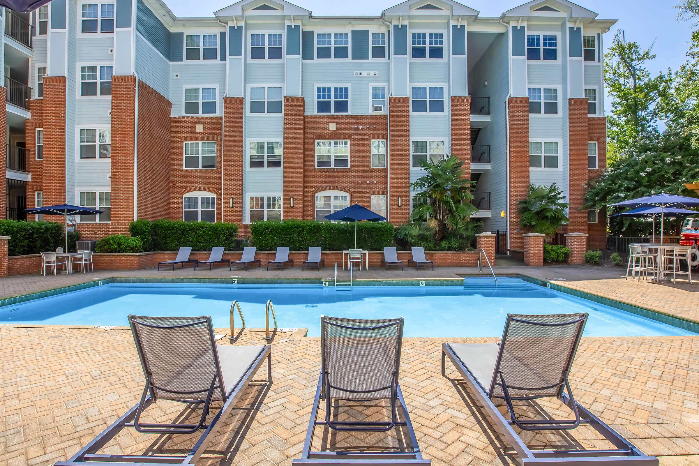 A view of an apartment complex with a swimming pool in the foreground. Three lounge chairs are positioned by the poolside, and several umbrellas provide shade. The building features brick and light blue exteriors, with lush greenery and a patio area visible in the background.