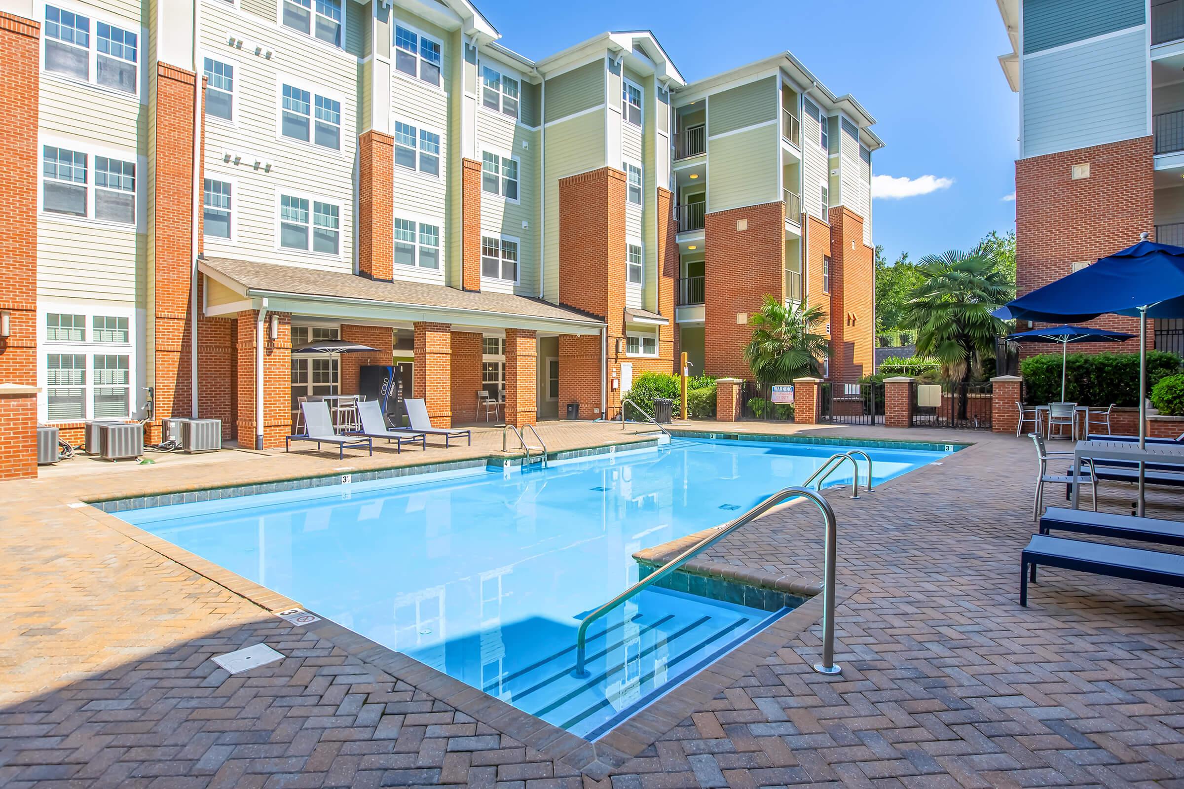 A spacious outdoor swimming pool surrounded by brick-paved flooring and lounge chairs. The pool area is flanked by modern apartment buildings with large windows. Lush greenery and palm trees are visible in the background, creating a relaxing atmosphere. Clear blue skies overhead.