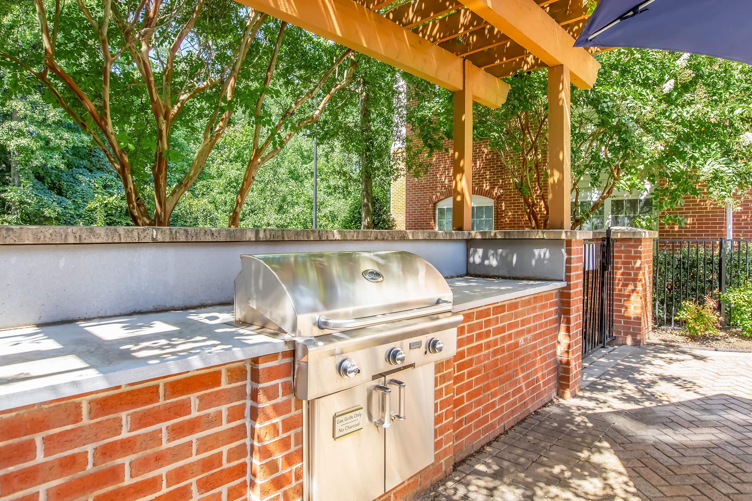 Stainless steel gas grill on a brick patio under a wooden pergola, surrounded by lush greenery. The grill features knobs and a clean surface, ideal for outdoor cooking. Sunlight filters through the trees, creating dappled shadows on the patio floor.