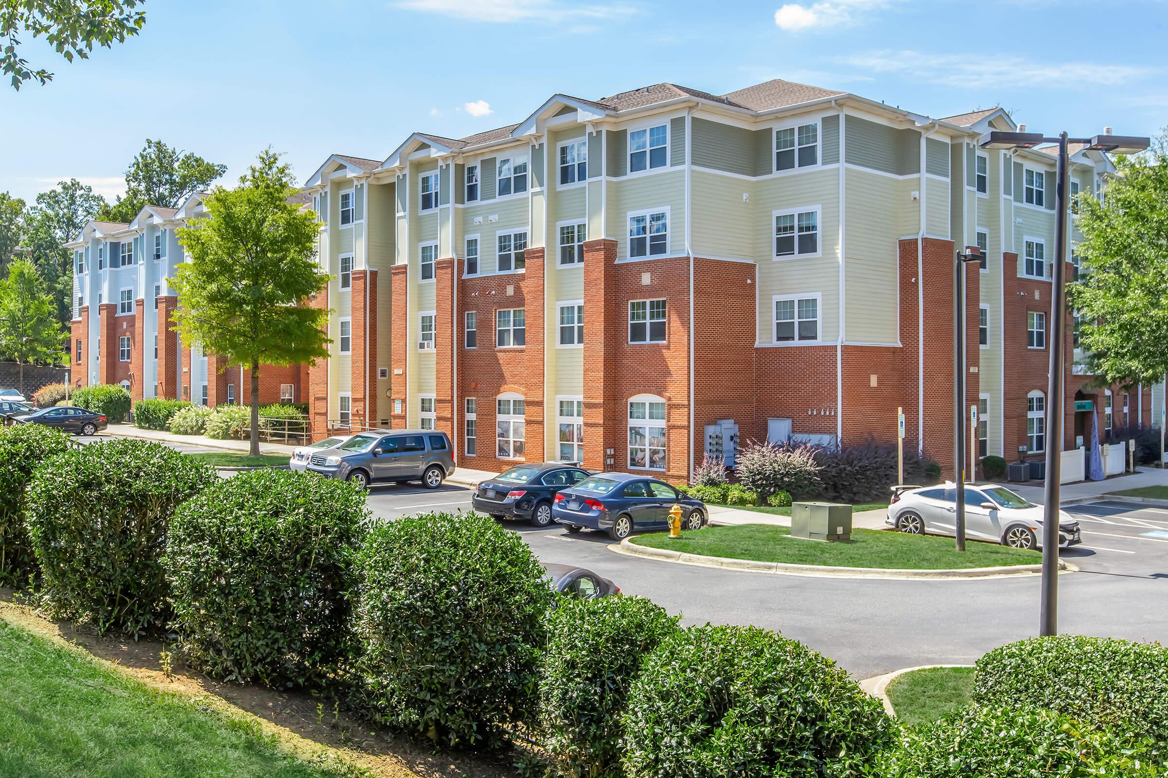 A modern apartment building featuring a mix of brick and siding exteriors. The three-story structure is set in a landscaped area with manicured shrubs and trees. Several parked cars are visible in front, and the sky is bright with a few clouds, indicating a sunny day.