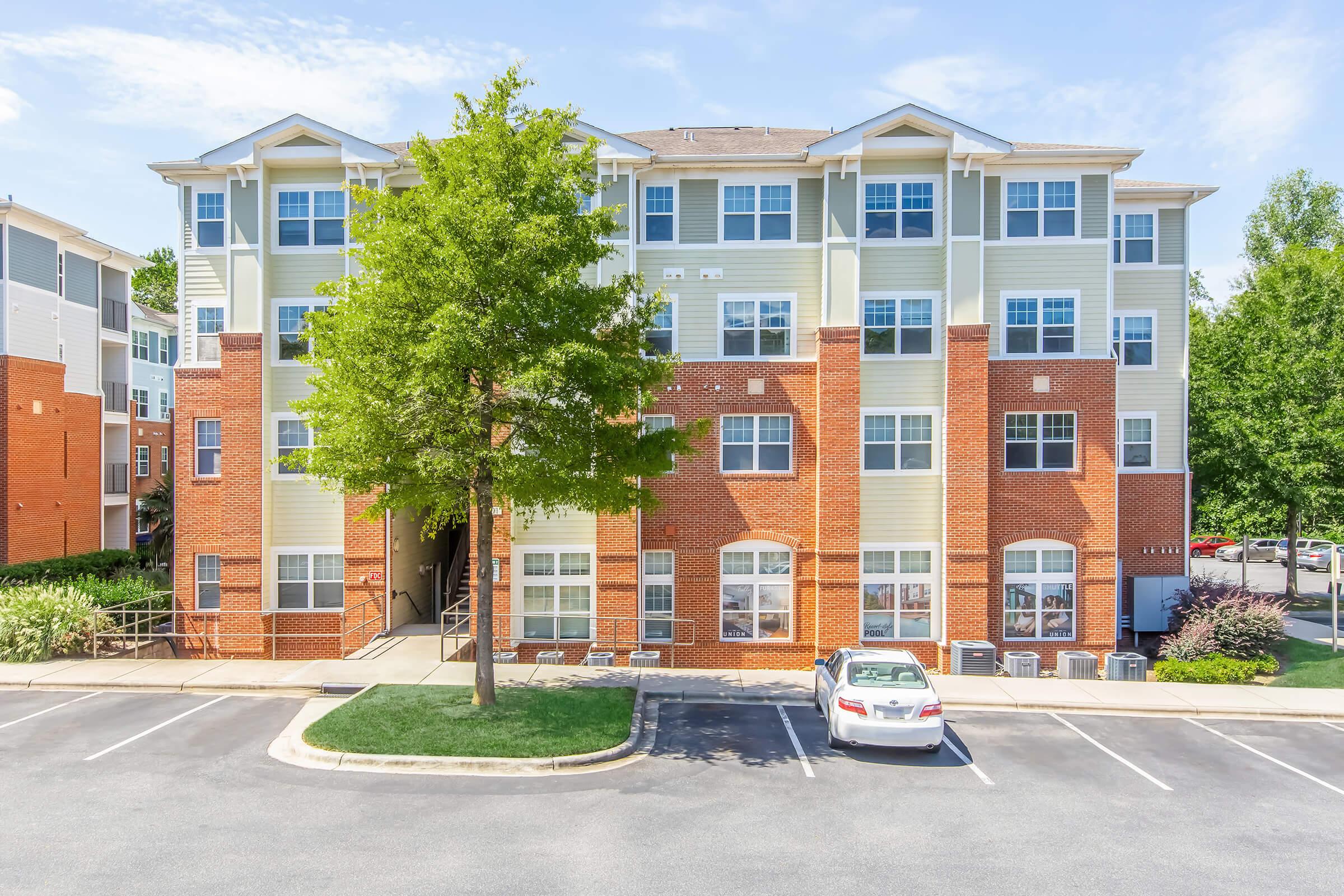 Four-story brick apartment building with light-colored siding and multiple windows. The ground floor features a covered entrance, surrounded by greenery and a parking lot with a white car parked in front. Clear blue sky and trees are visible in the background.