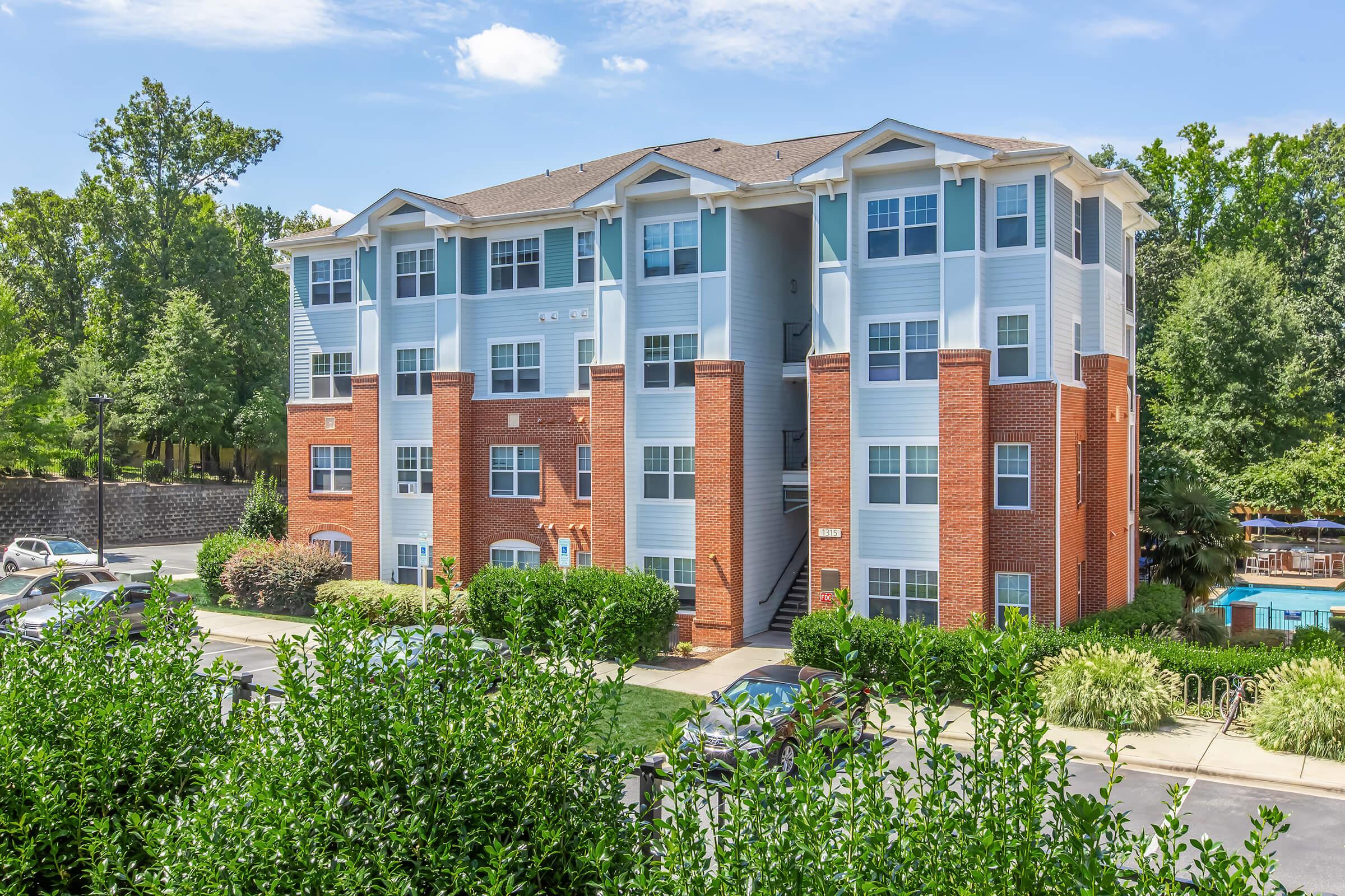 A modern, three-story apartment building featuring a mix of light blue siding and red brick. The building is surrounded by green landscaping, with a view of a swimming pool in the background. The sky is bright and partly cloudy, creating a welcoming atmosphere.