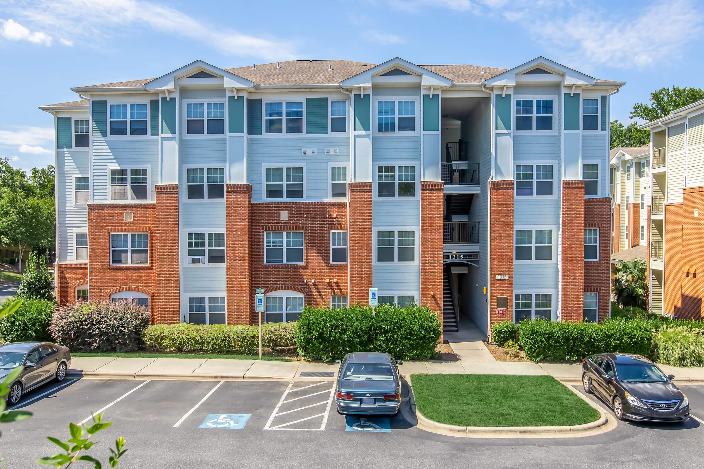 A multistory apartment building with a mix of brick and blue siding. The building features several windows, balconies, and a landscaped area in front with parking spaces visible. Clear blue sky and greenery surround the scene, creating a welcoming environment.