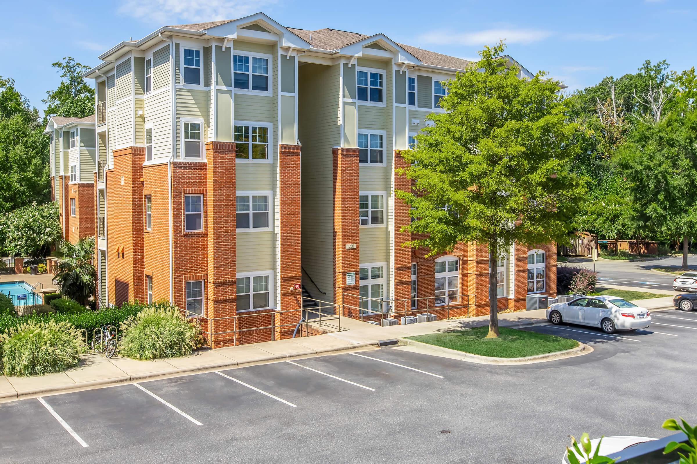 A three-story residential building with a mix of brick and siding exterior, surrounded by greenery. A sidewalk leads to the entrance, and a parking lot is in front with a few parked cars. The scene is bright and captures a pleasant, sunny day.