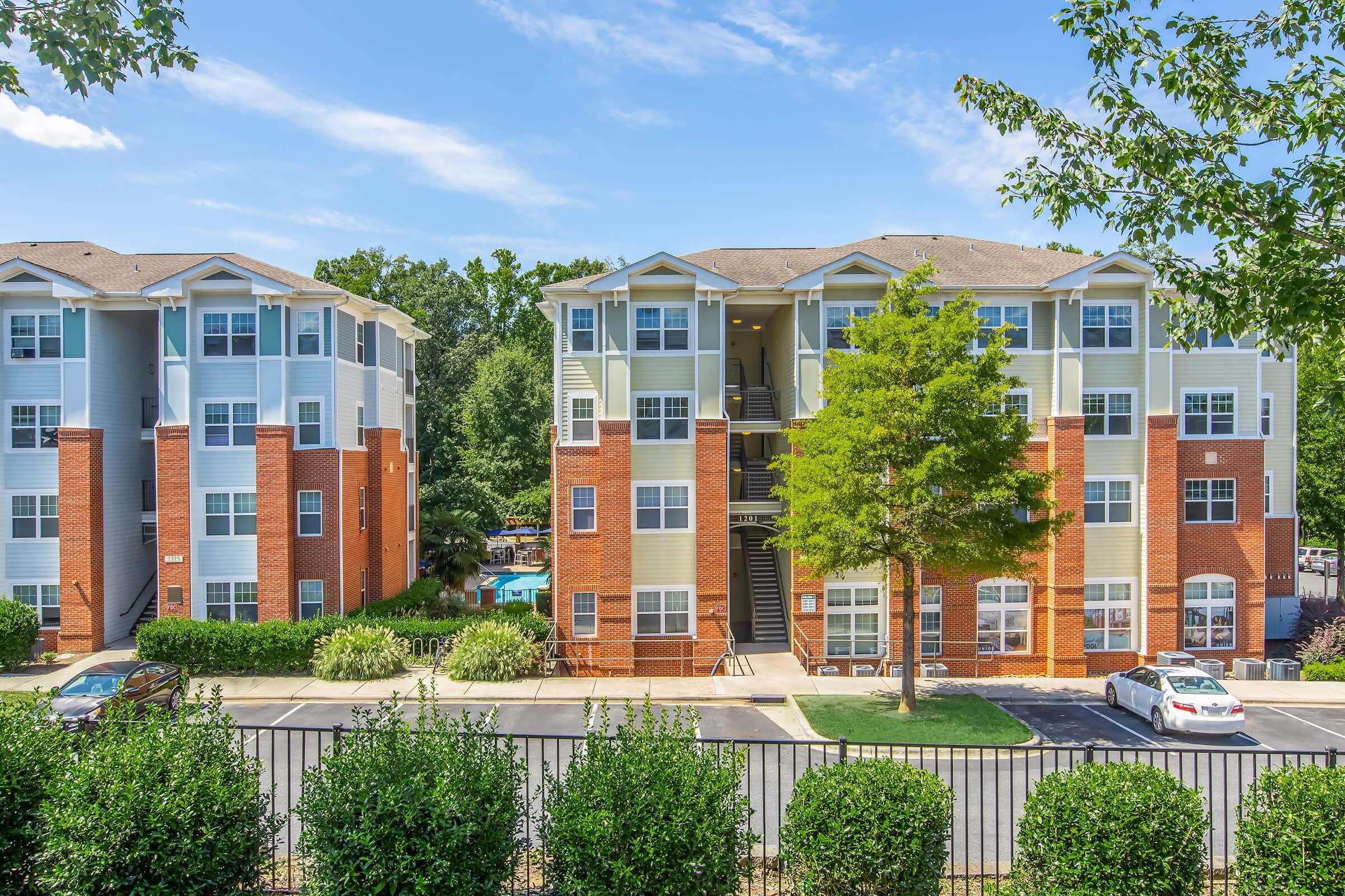 A view of two residential apartment buildings. The buildings are three stories tall, featuring a mix of brick and light-colored siding. In the foreground, there are manicured hedges and a parking lot with a couple of cars. The background includes trees and a clear blue sky.