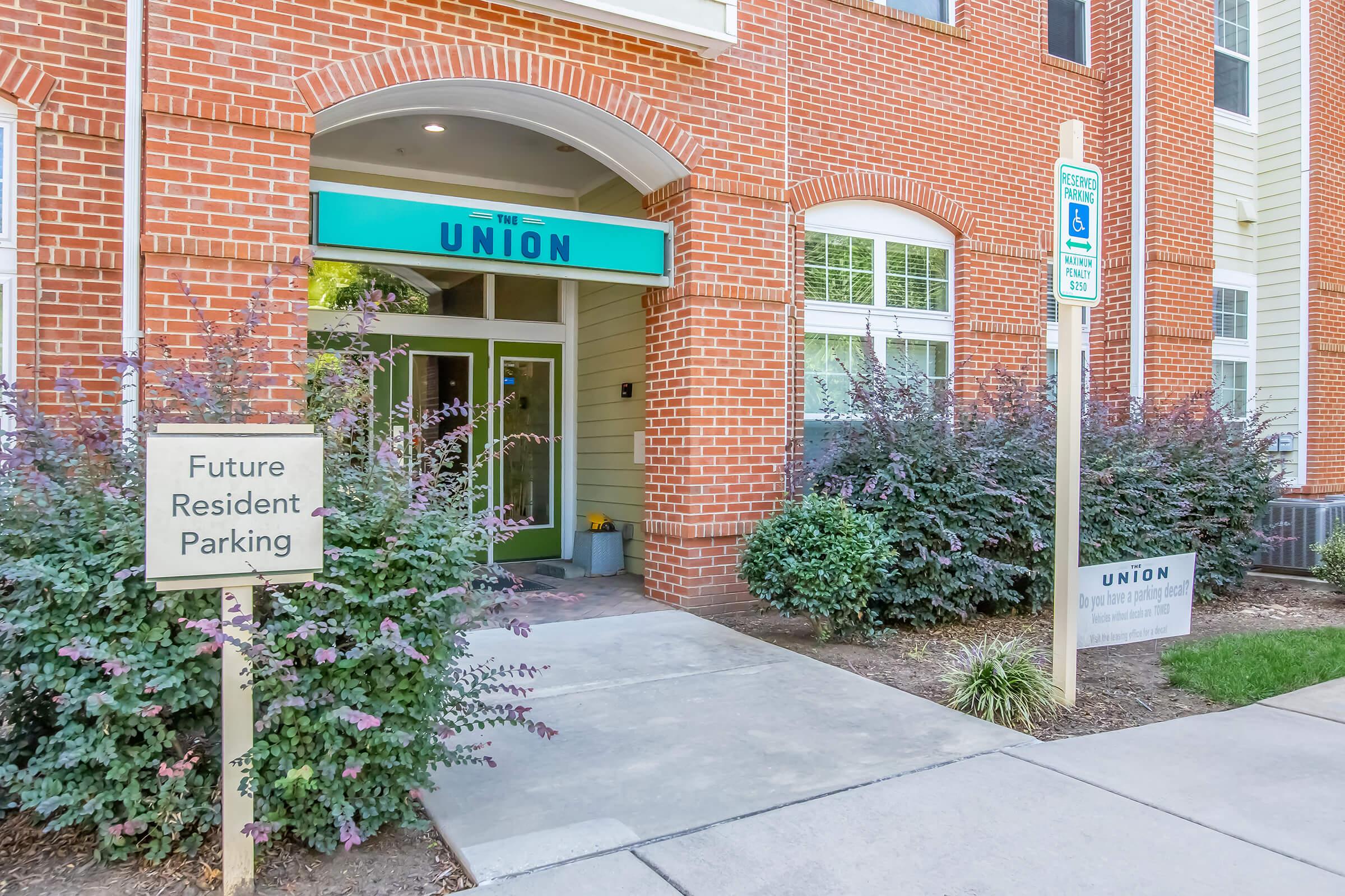 The entrance of a residential building called "The Union," featuring a green awning, glass doors, and brick walls. Nearby, there are signs indicating "Future Resident Parking" and "Union." Lush plants and shrubs frame the pathway leading to the entrance.