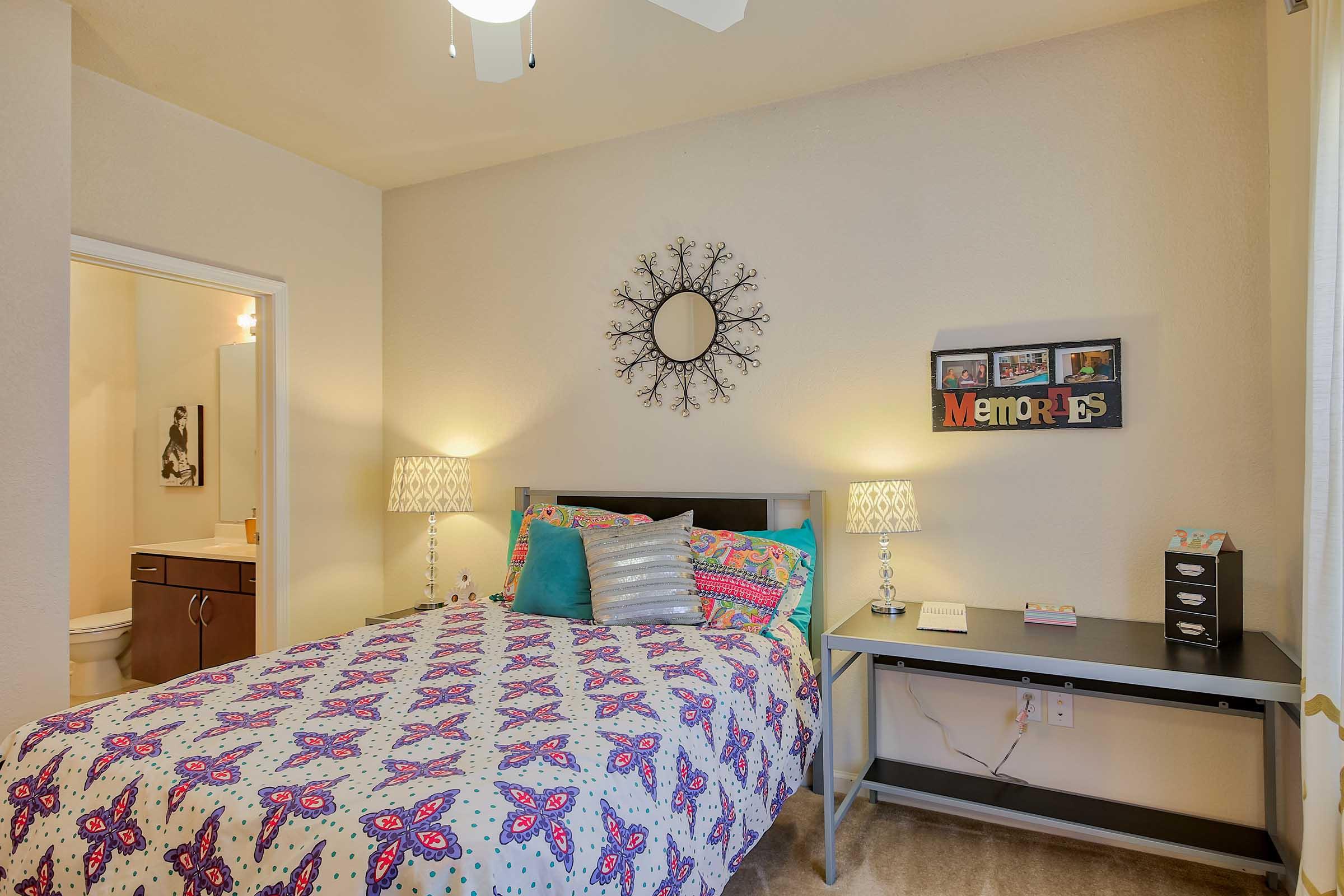 A cozy bedroom featuring a queen-sized bed with a colorful patterned duvet cover. Two bedside lamps provide soft lighting. A decorative wall mirror is above the bed, and a desk with storage stands nearby. A bathroom is visible through an open door in the background. Warm, inviting decor throughout.