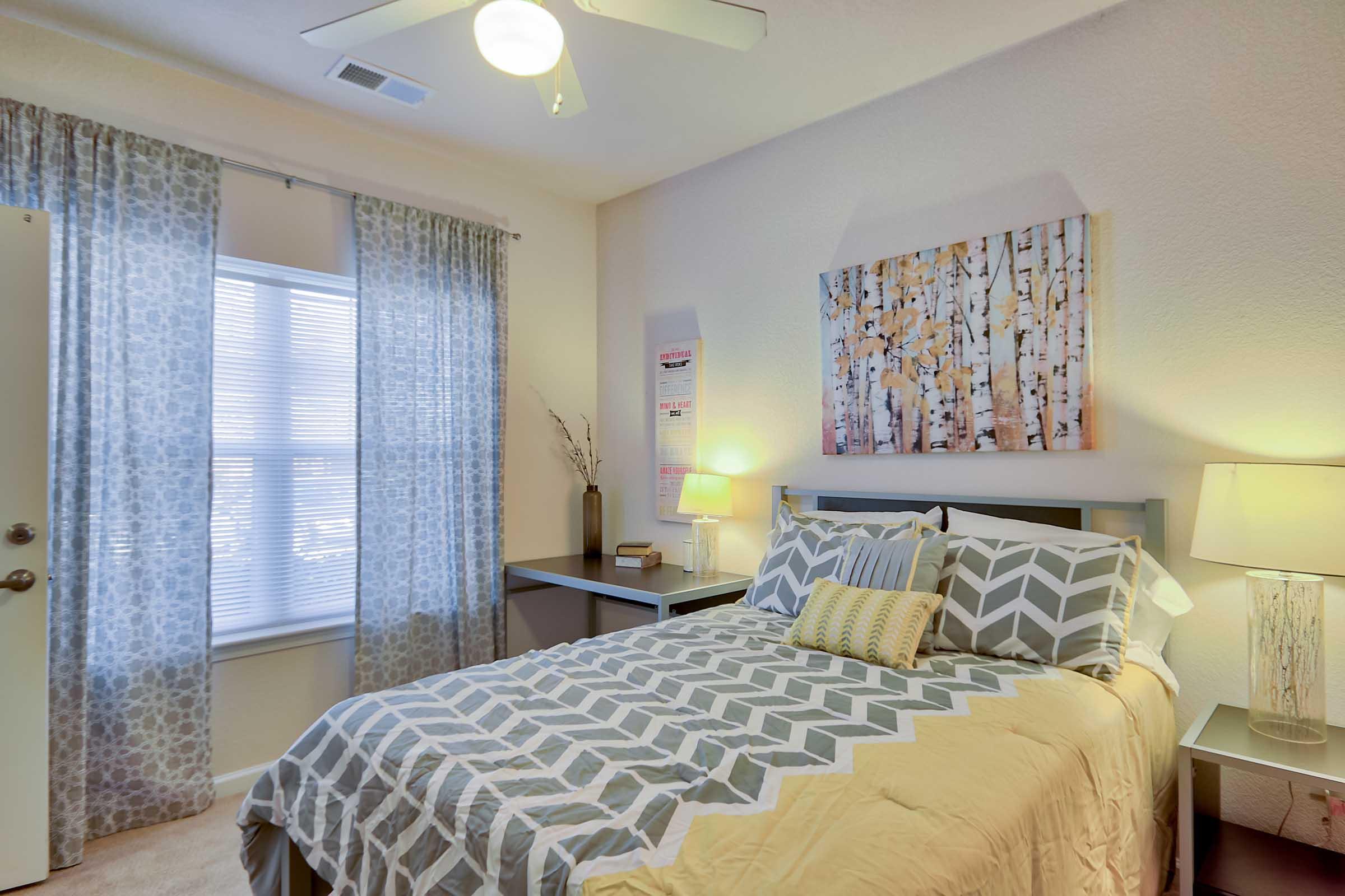 A cozy bedroom featuring a queen-sized bed with a patterned gray and yellow quilt. There are two lamps beside the bed, a wooden desk with a chair, and large windows with sheer curtain drapes. An abstract artwork of birch trees hangs above the bed, creating a warm and inviting atmosphere.