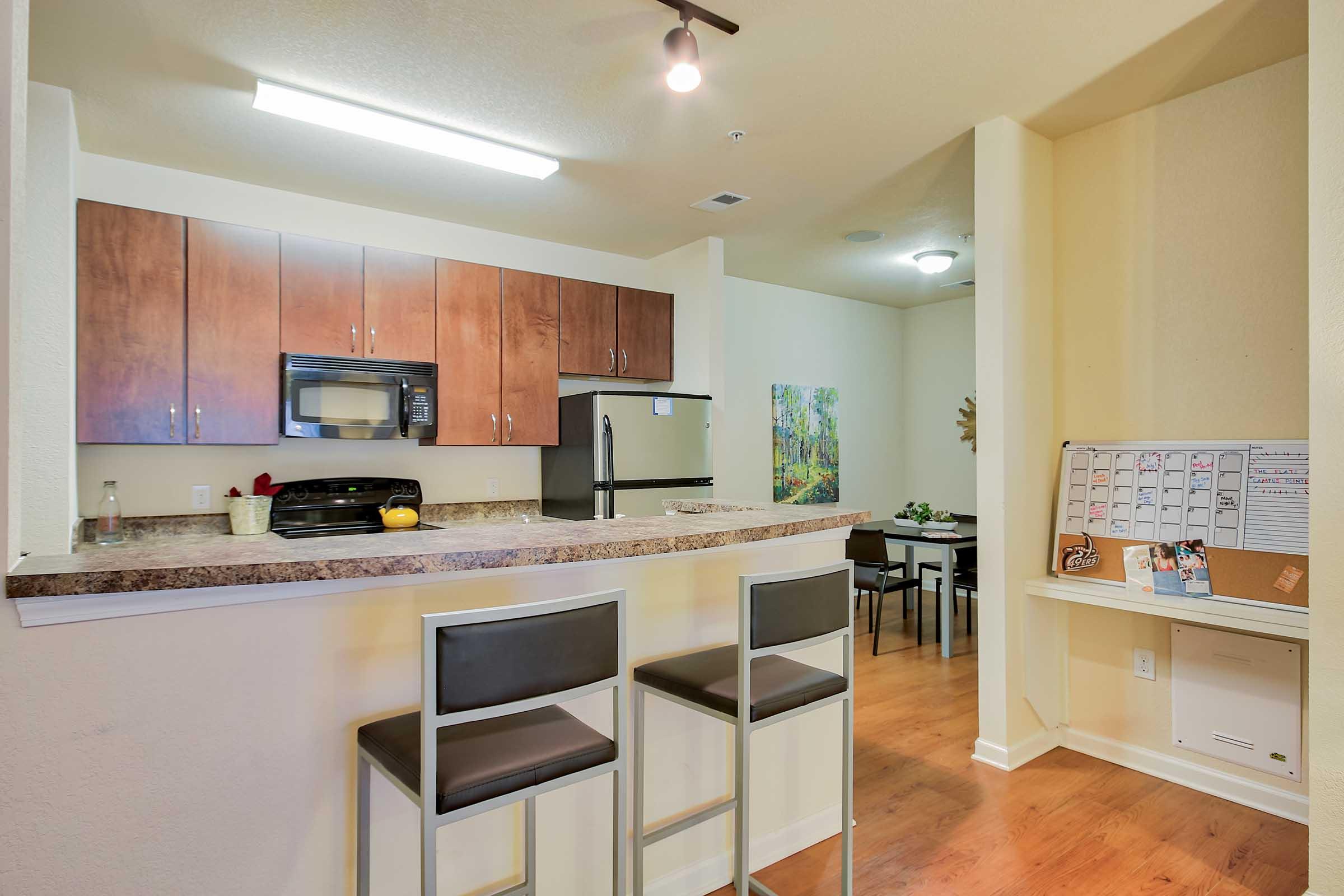 Modern kitchen area featuring a bar with two high stools, brown cabinetry, and stainless steel appliances. A large window provides natural light, while a dining area with a table and chairs is visible in the background. The space is clean, bright, and well-organized.