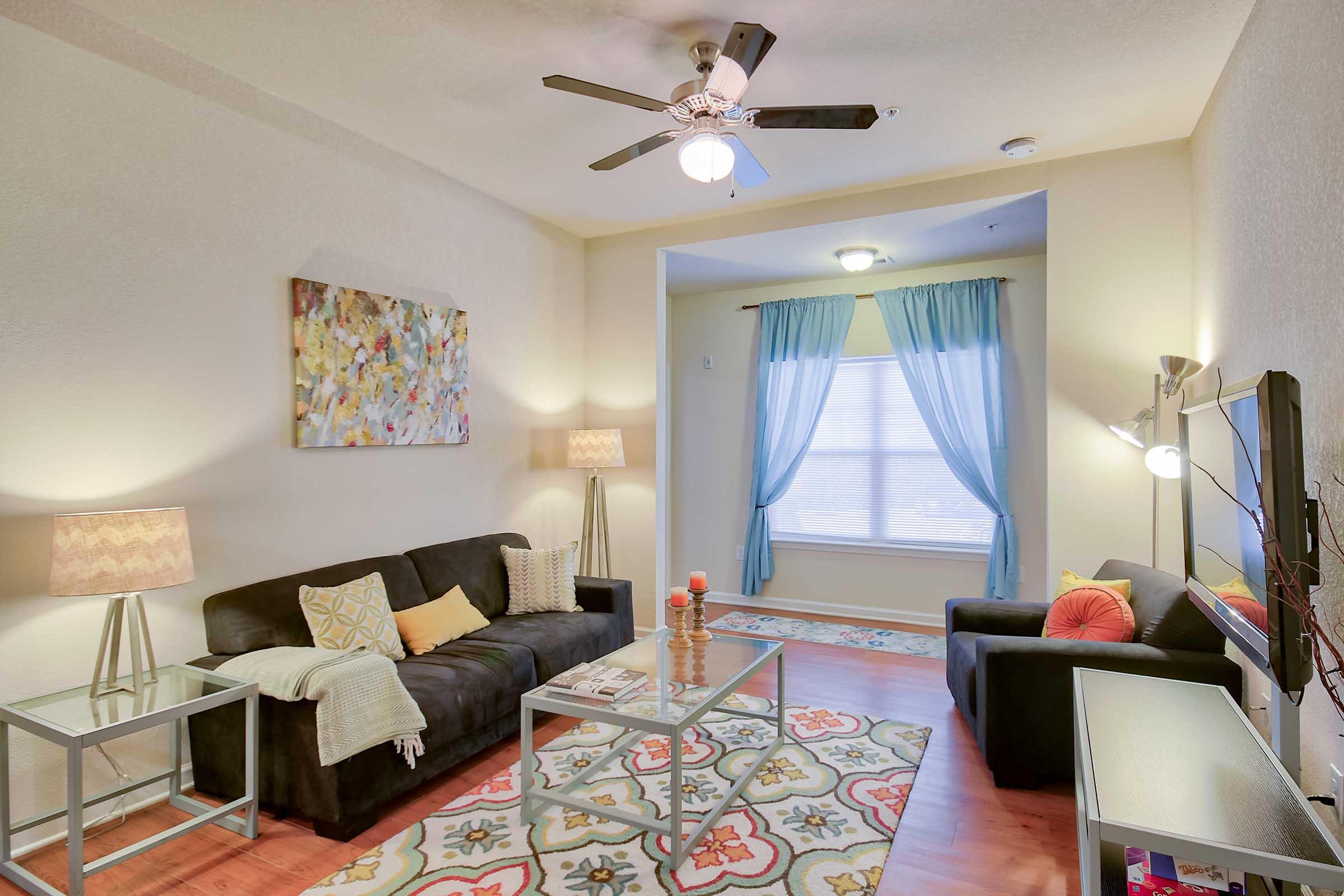 A cozy living room featuring two dark sofas with colorful cushions, a coffee table with magazines, and a patterned area rug. Natural light filters through sheer curtains by the window, complemented by a ceiling fan and stylish lamps. Wall art adds a touch of color to the space.