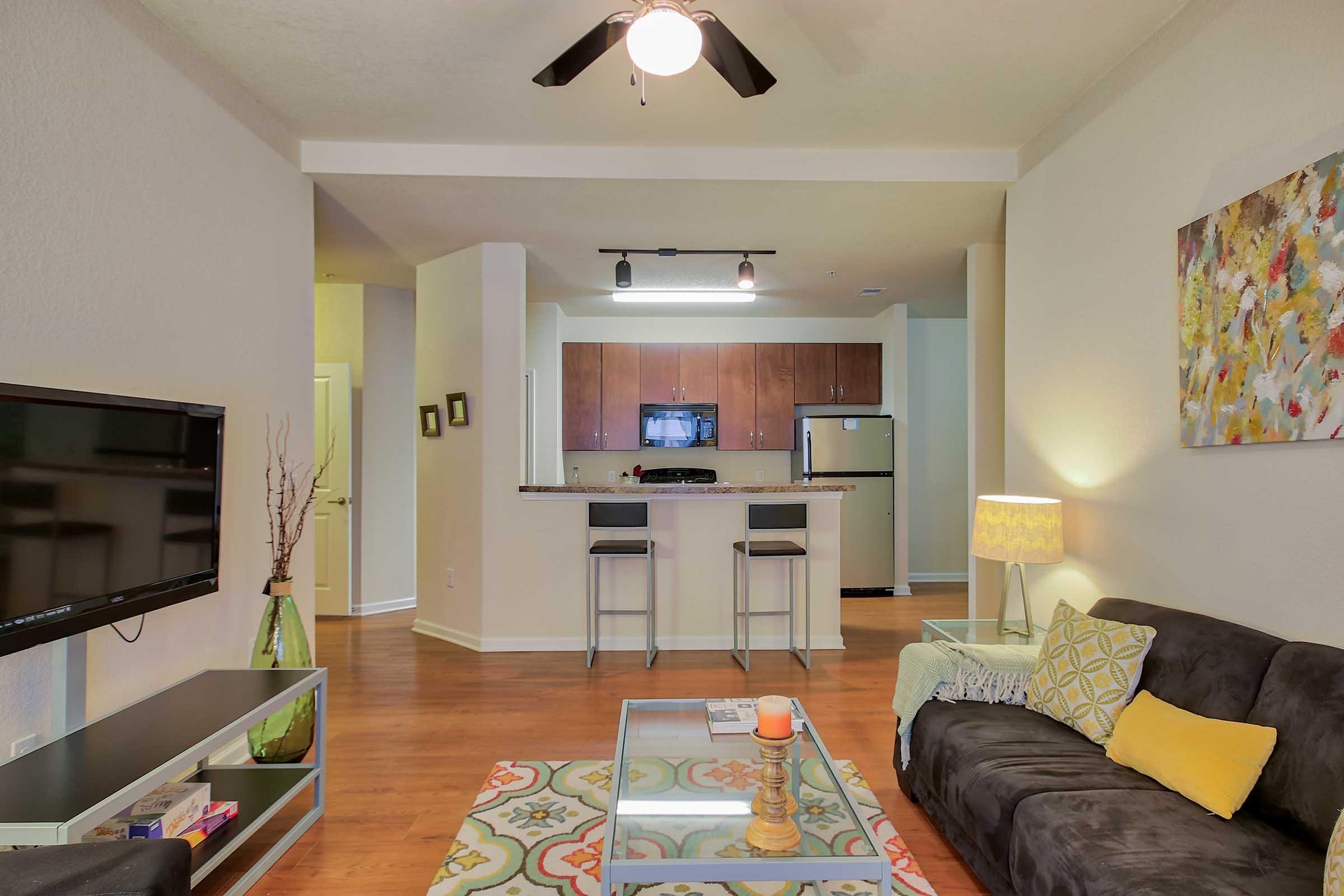 A modern living room featuring a dark sofa, colorful patterned rug, and a coffee table. In the background, a dining area with two bar stools and a kitchen visible, showcasing wooden cabinets and stainless steel appliances. Decorative lamp and artwork add warmth to the space.