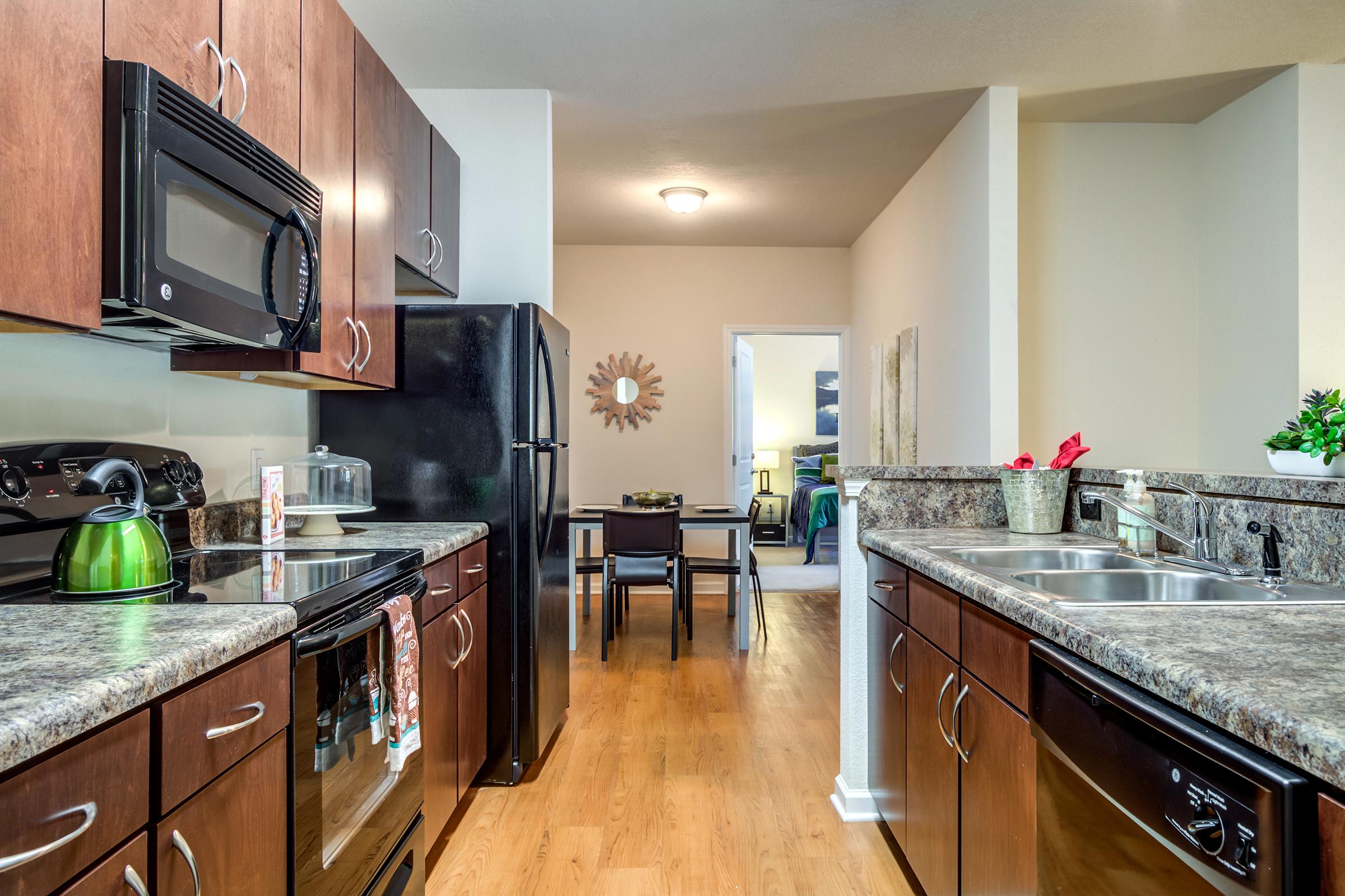 A modern kitchen featuring dark wood cabinets, granite countertops, and stainless steel appliances. There's a microwave, stove, and refrigerator, along with a kettle on the counter. In the background, a dining area is visible with a table and chairs, and an adjoining living space beyond.