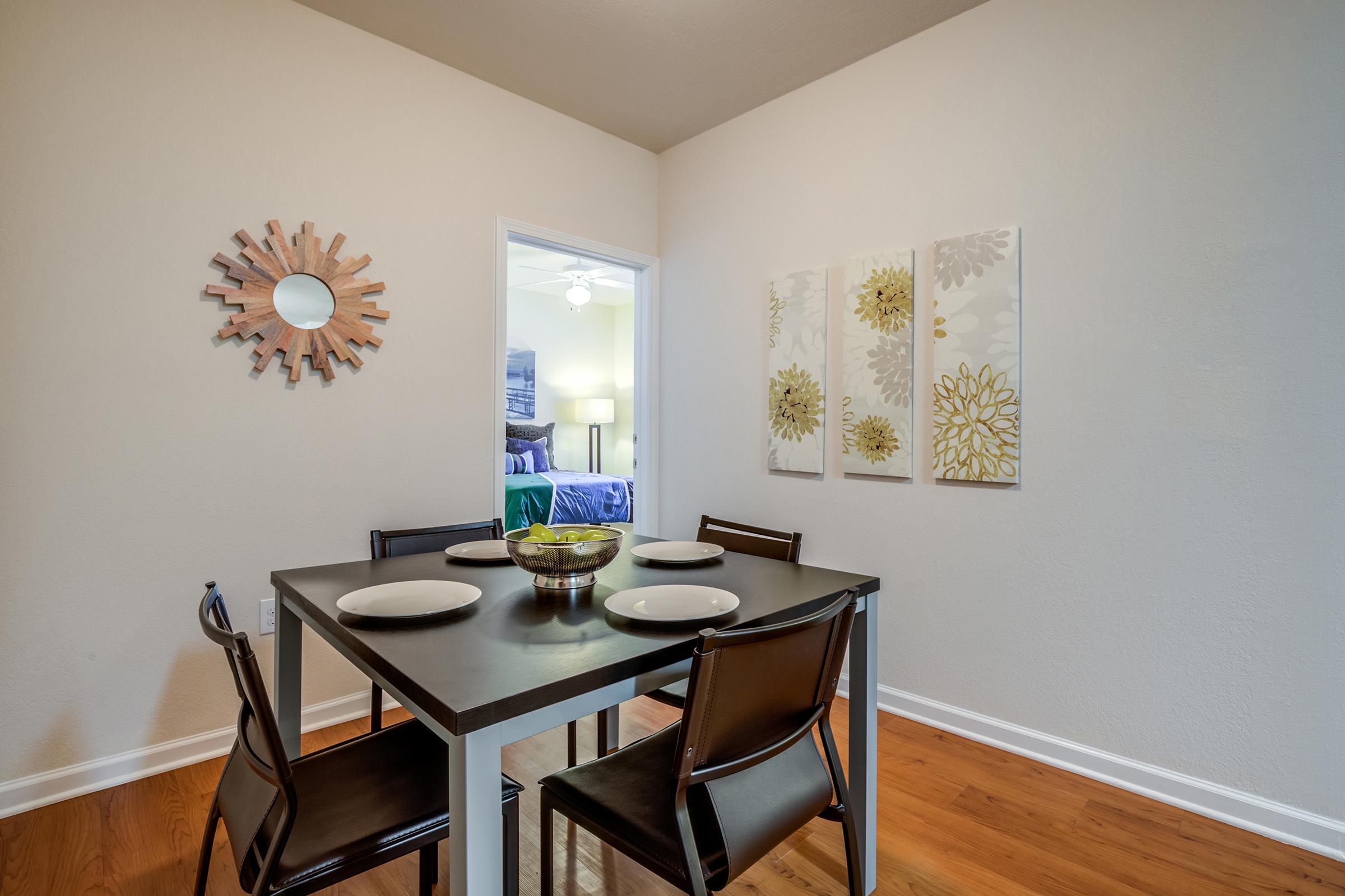 A modern dining area featuring a square black table set with white plates and a bowl of fruit, surrounded by four black chairs. On the wall, there's a decorative wooden mirror and three floral artwork pieces. A doorway in the background leads to a brightly lit room with a bed and a ceiling fan.