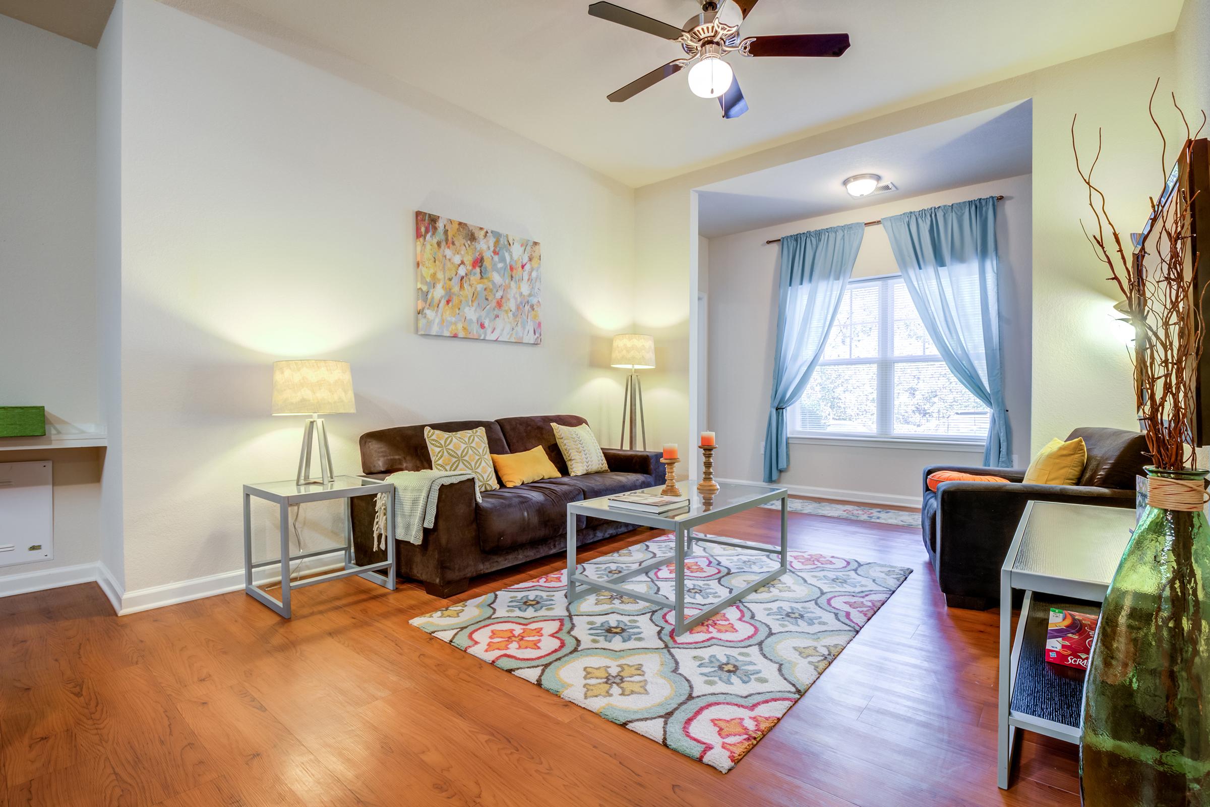 A cozy living room featuring a brown couch and vibrant decorative pillows. A glass coffee table sits on a colorful patterned rug, surrounded by two lamps with warm light. A large window is dressed with light blue curtains, allowing natural light to brighten the space. A decorative vase and a piece of wall art enhance the decor.
