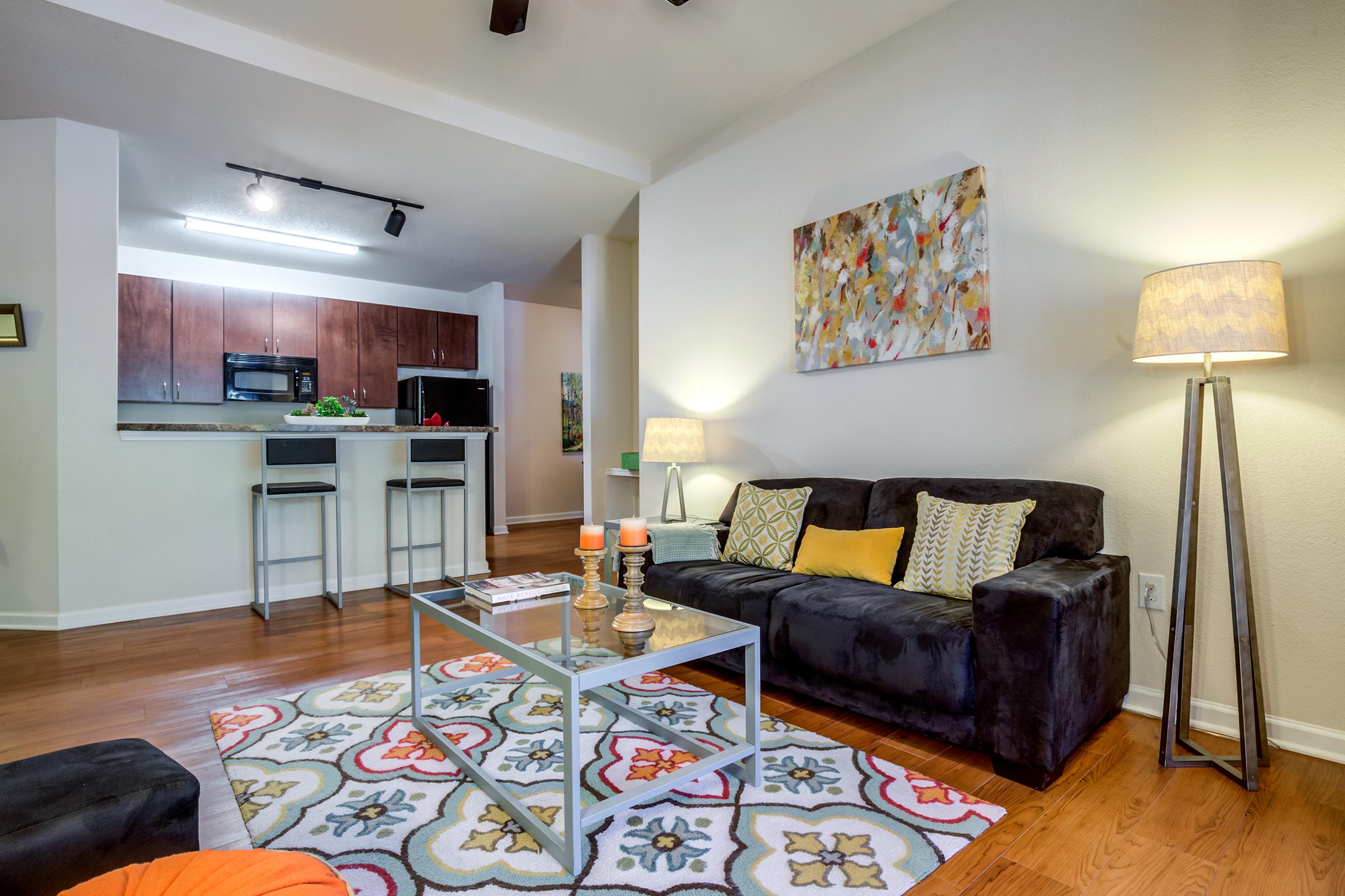 A cozy living room featuring a dark sofa with colorful cushions, a glass coffee table, and a patterned area rug. A large abstract painting decorates the wall, and there are two lamps providing warm light. In the background, a kitchen with dark cabinetry and modern appliances is visible.