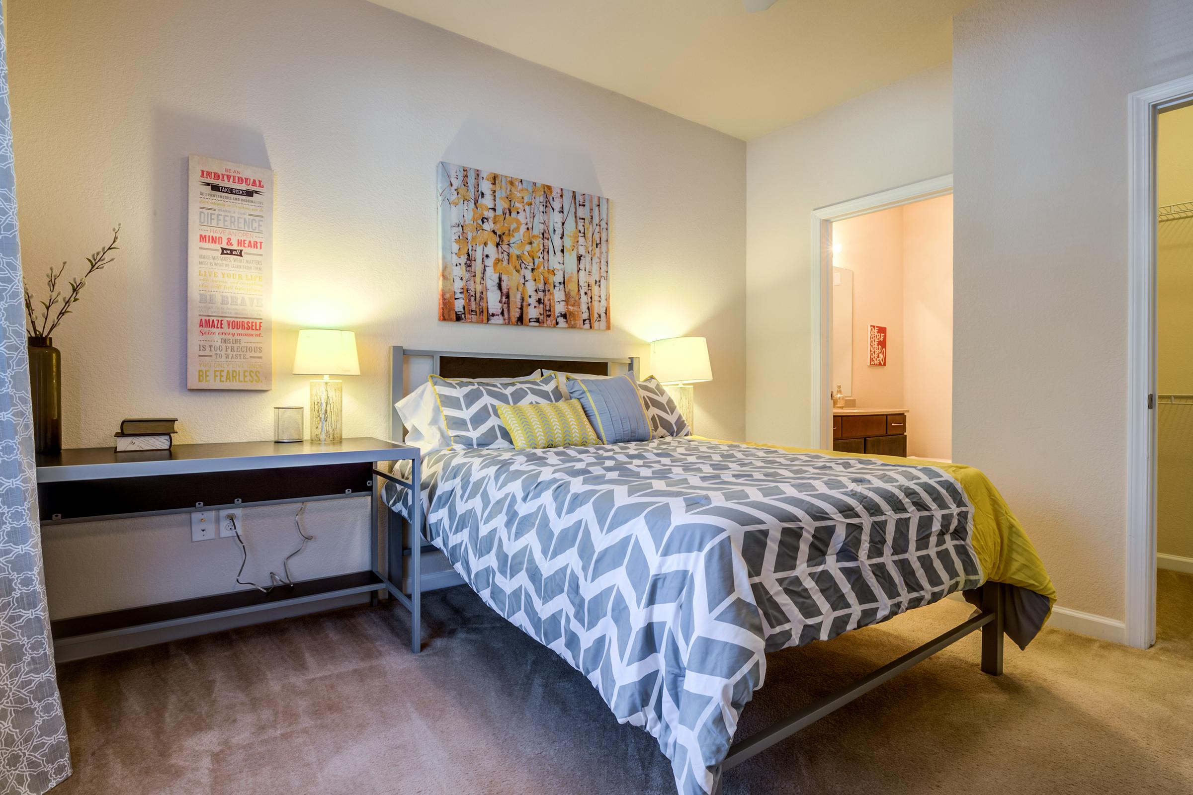 A cozy bedroom featuring a bed with a patterned duvet and decorative pillows. There are two bedside lamps, a small desk with books, and a piece of artwork on the wall. The room has light-colored walls and carpet, creating a warm and inviting atmosphere. A doorway leads to a bathroom in the background.