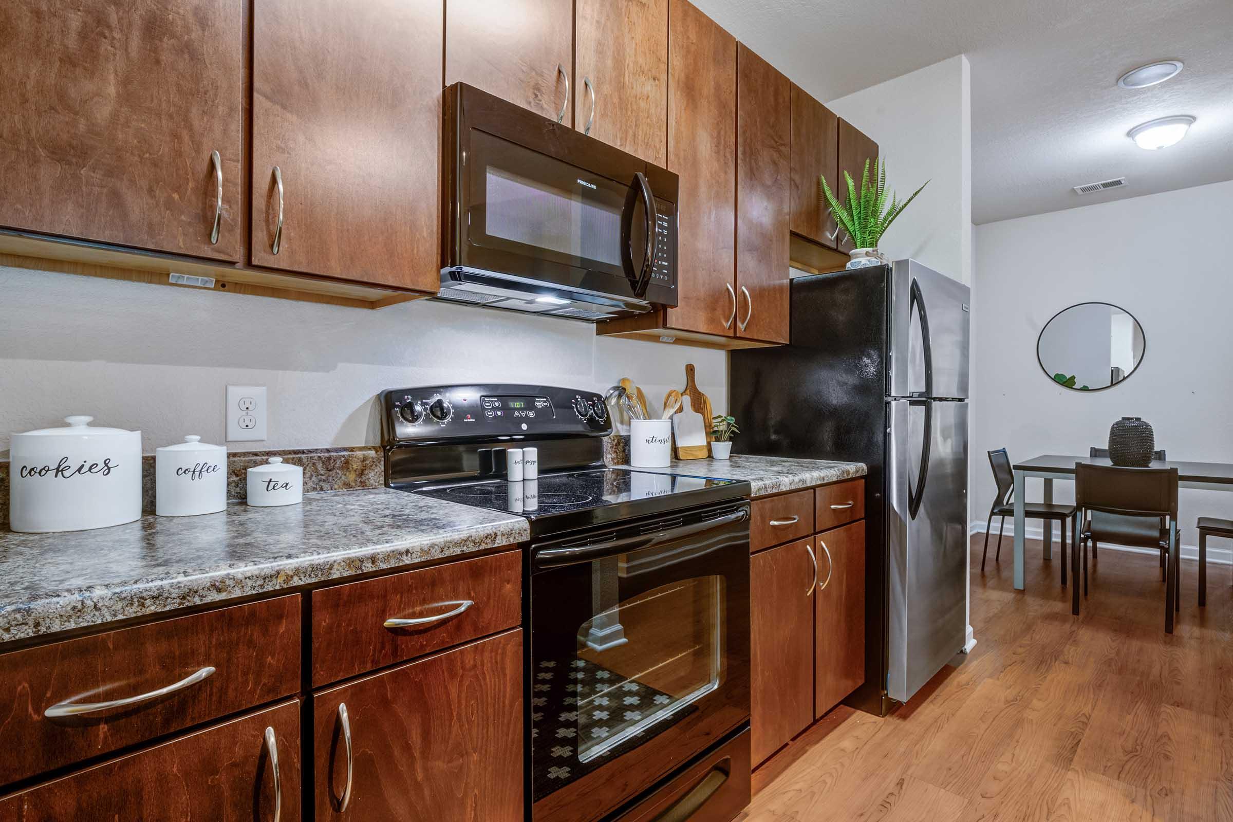A modern kitchen featuring dark wood cabinetry, a granite countertop, and stainless steel appliances. There is a black oven and microwave, along with decorative canisters labeled "cookies," "coffee," and "tea." A dining area with a round mirror and a small table is visible in the background.