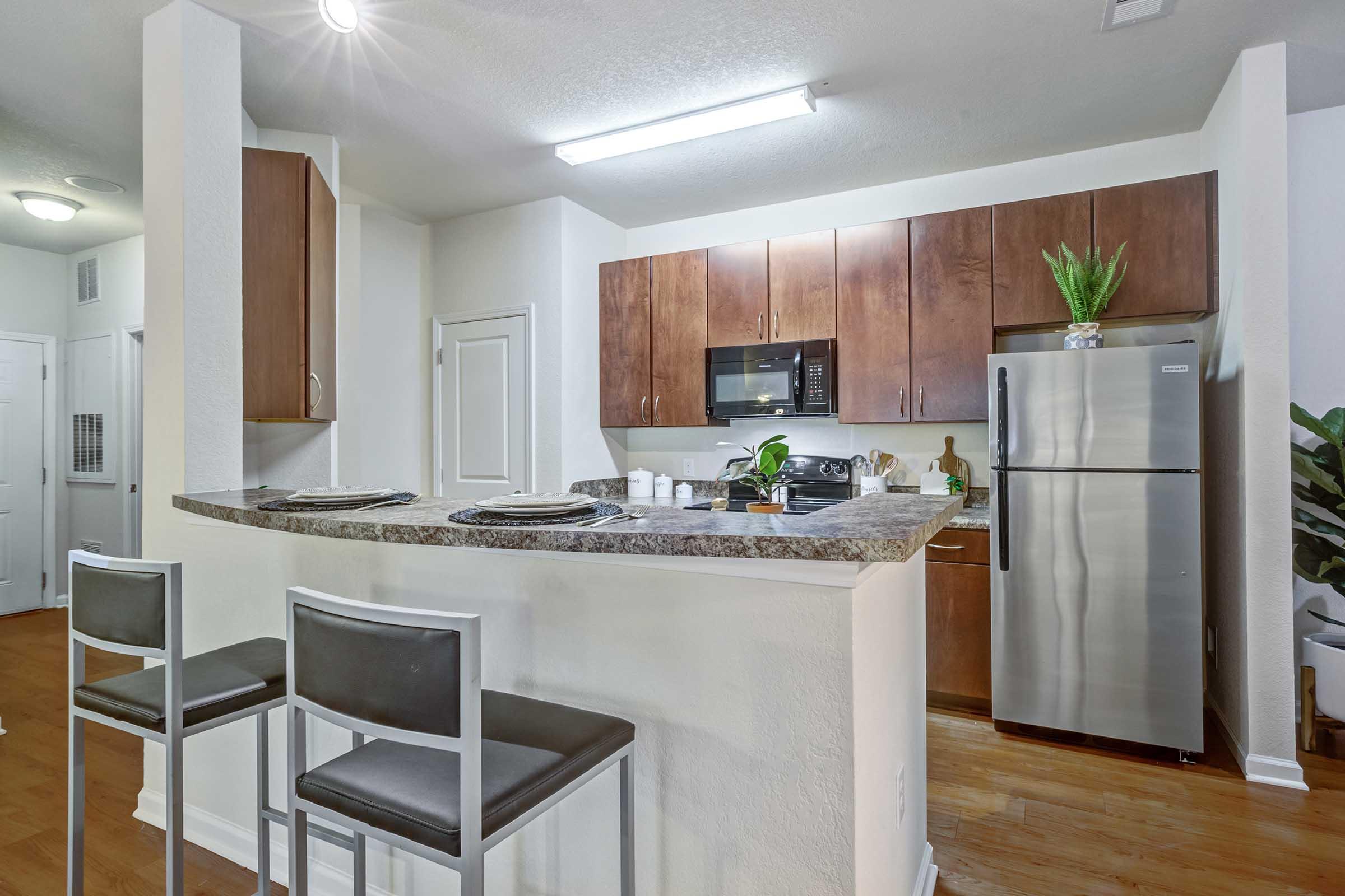 Modern kitchen featuring wooden cabinets, stainless steel appliances, and a granite countertops. Two bar stools are positioned at a breakfast bar. The kitchen is bright with overhead lighting, and there are decorative plants on the countertop. The space has a clean and contemporary design.