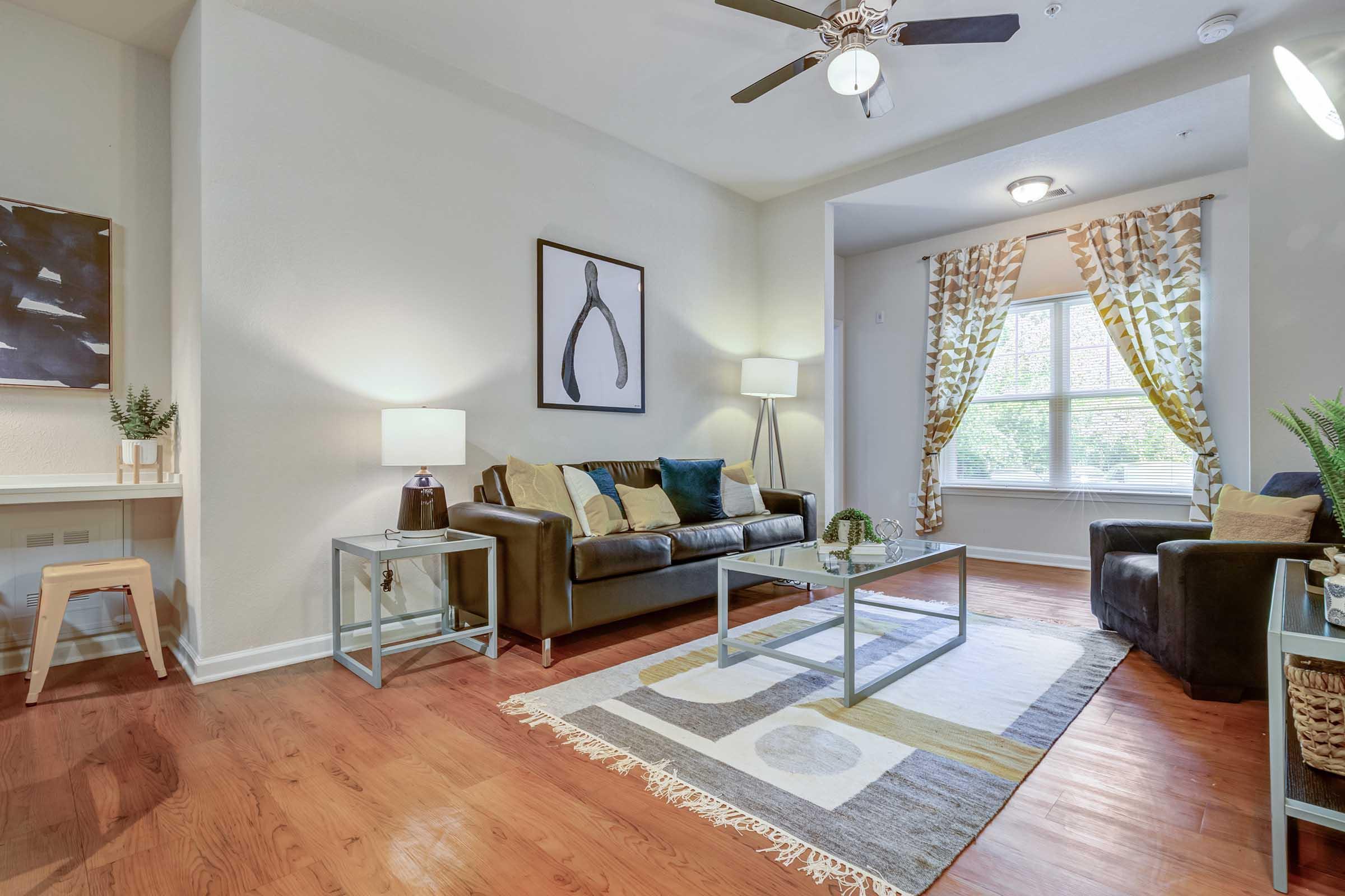 Modern living room featuring a brown leather sofa with colorful cushions, a glass coffee table on a patterned rug, and light wood flooring. There are two side tables with lamps, a window adorned with patterned curtains, and a small desk with a chair in the background. Decor includes art on the wall and a plant.