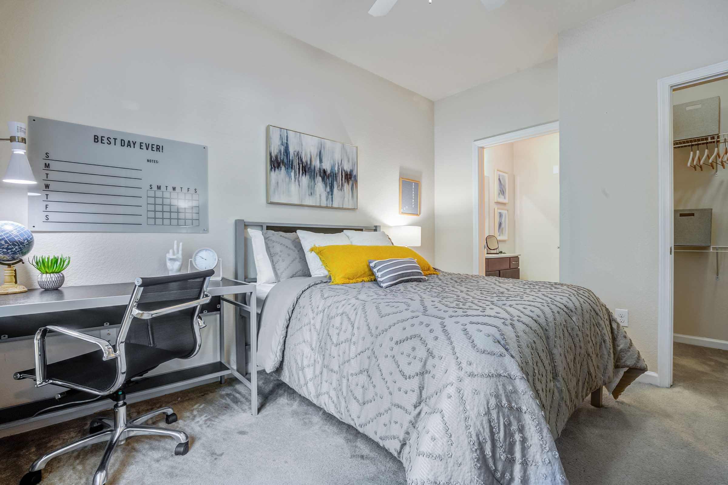 A well-decorated bedroom featuring a bed with gray and yellow bedding, a stylish desk with a globe and plant, and a wall calendar saying "BEST DAY EVER!" A closet is visible in the background, and the room has a light, airy feel with neutral colors.