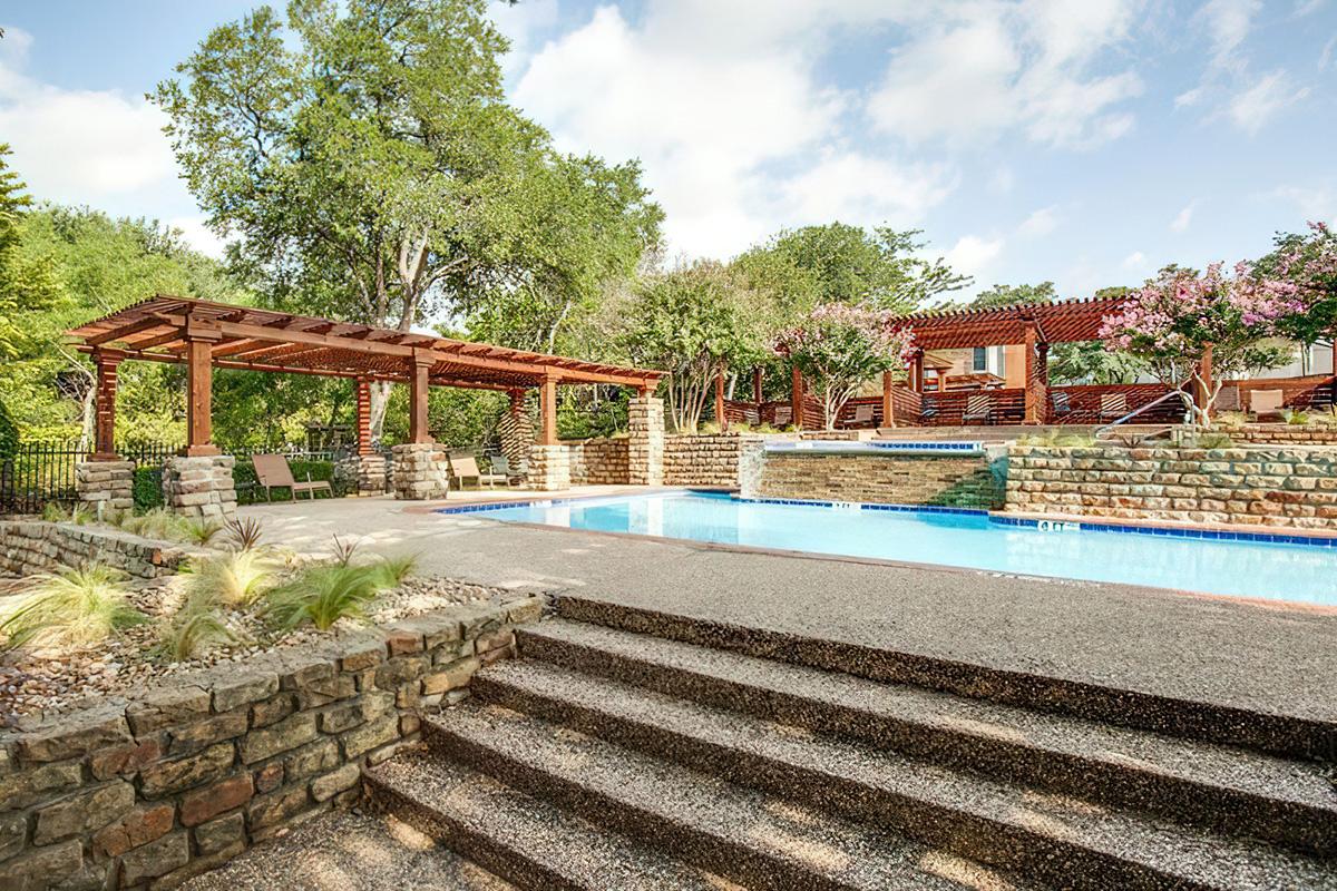 A serene outdoor pool area featuring a crystal-clear swimming pool surrounded by stone walls and steps. There are wooden pergolas shaded by lush trees, with lounge chairs placed nearby, and flowering plants adding color to the landscape. Bright blue skies and fluffy clouds create a tranquil atmosphere.